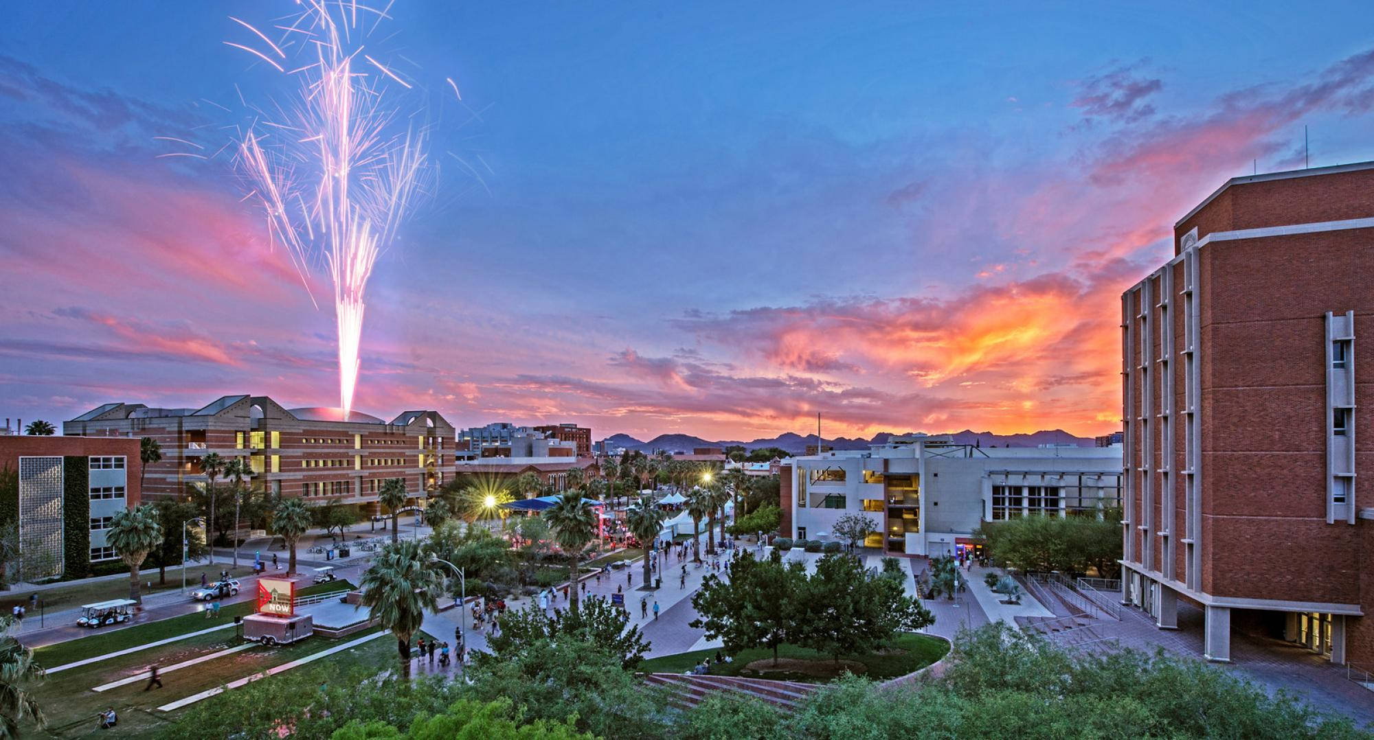 Fireworks 桌布 壁紙 — 煙火在黃昏時分的大學校園上空綻放，將粉橙色光芒投射在磚砌建築物、棕櫚樹和行走的學生身上。