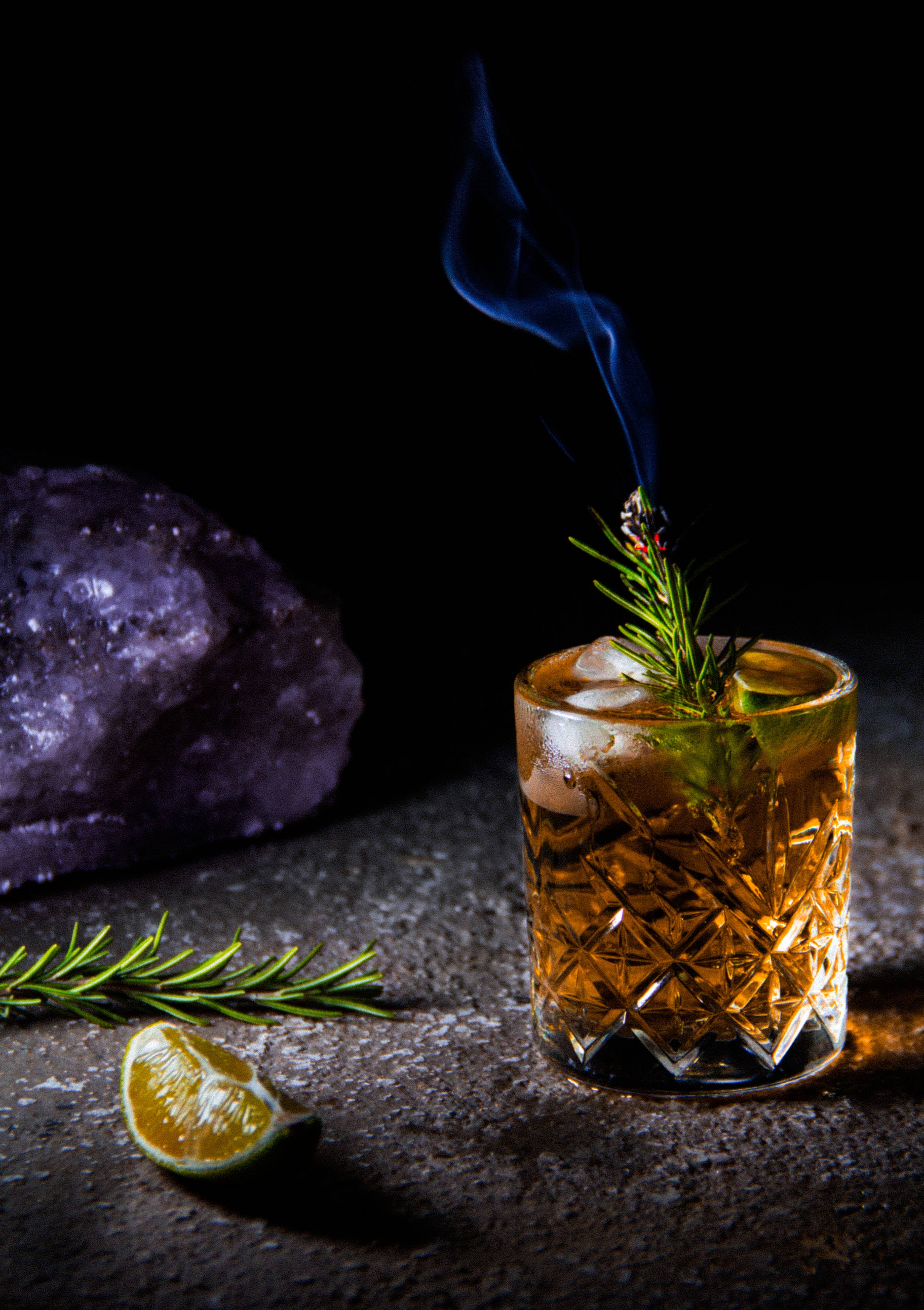 alcohol 桌布 壁紙 — 煙燻琥珀色雞尾酒盛載於雕花玻璃杯中，配以迷迭香和柑橘，在深色背景下於紫色水晶旁閃耀光芒。