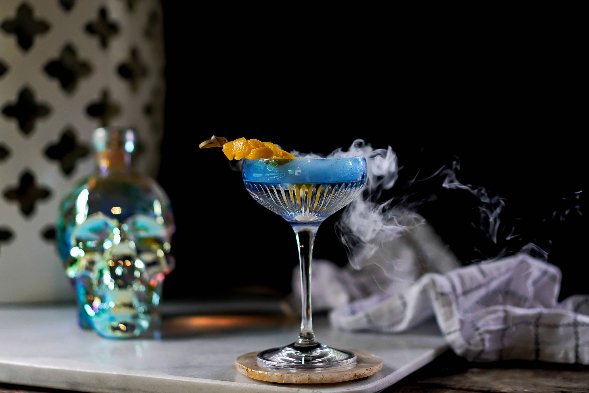 Drink 桌布 壁紙 — 煙霧藍色雞尾酒盛載於水晶碟形杯，飾以柑橘和骷髏形狀的瓶子，在黑暗中發光。