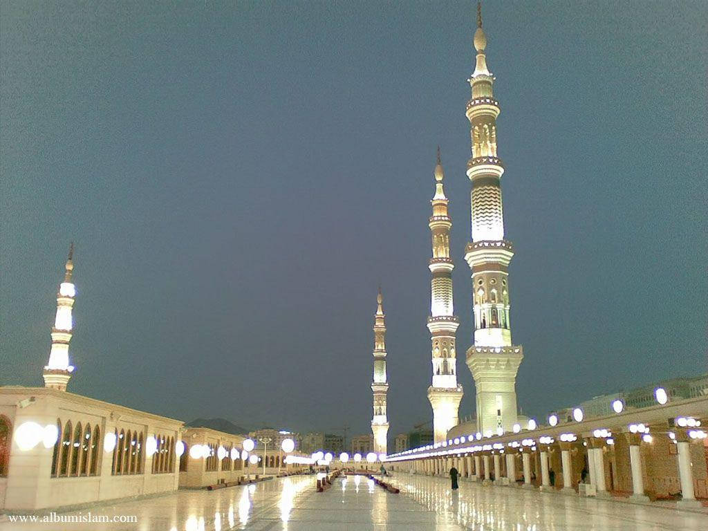 Madina 桌布 壁紙 — 照亮了的清真寺宣禮塔在暮色天空下升起，倒映在濕潤的庭院路面上。