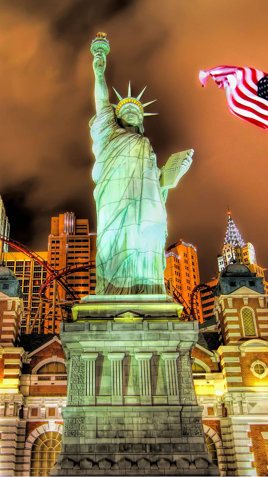 Vegas iPhone 桌布 壁紙 — 照亮嘅綠色自由女神像喺溫暖嘅橙色夜空下高高聳立，背後有飄揚嘅美國國旗同城市天際線。