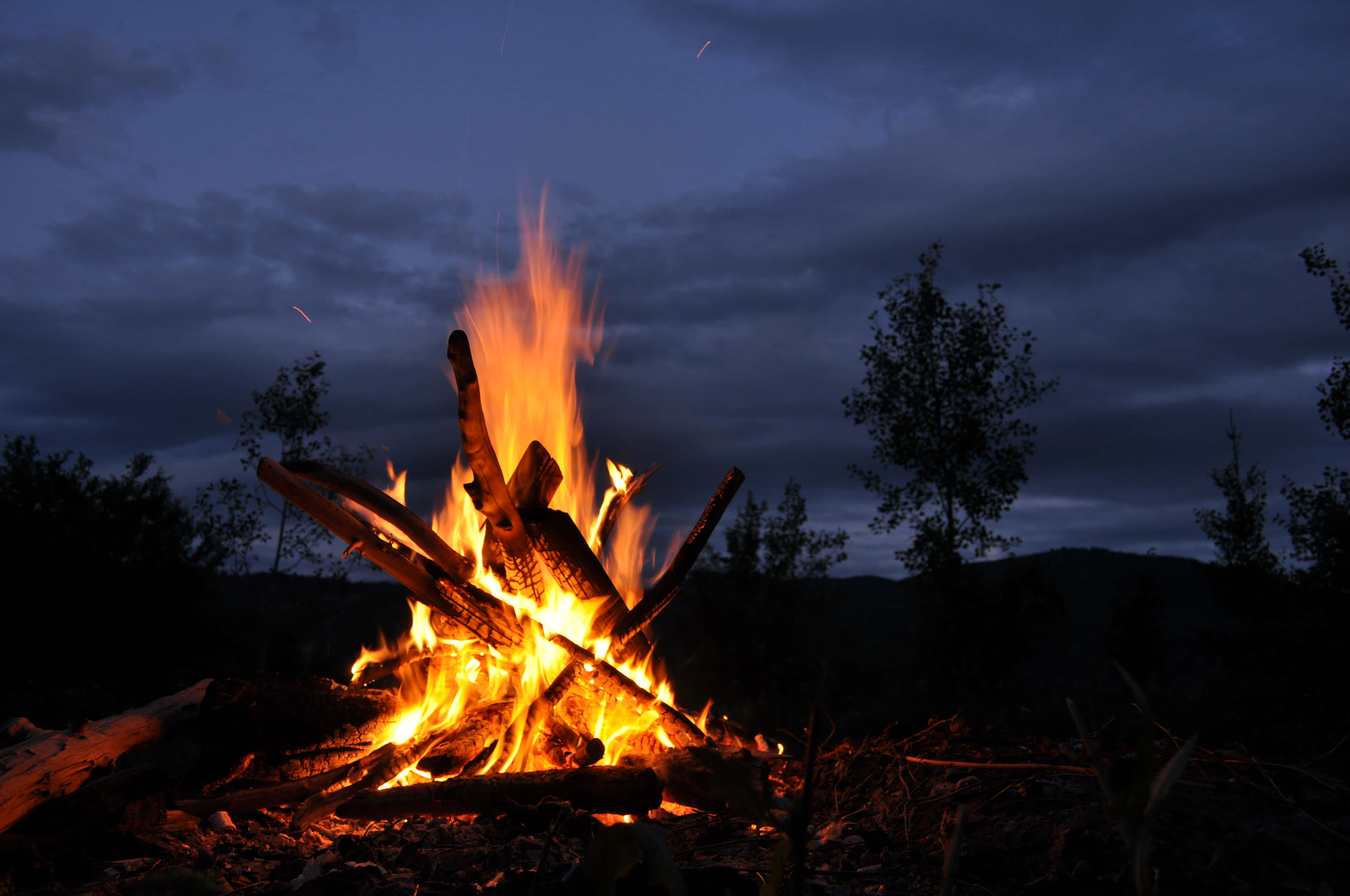 Bonfire 桌布 壁紙 — 熊熊營火在暮色蒼茫的天空下明亮地燃燒，襯托著樹木和遠山的剪影。