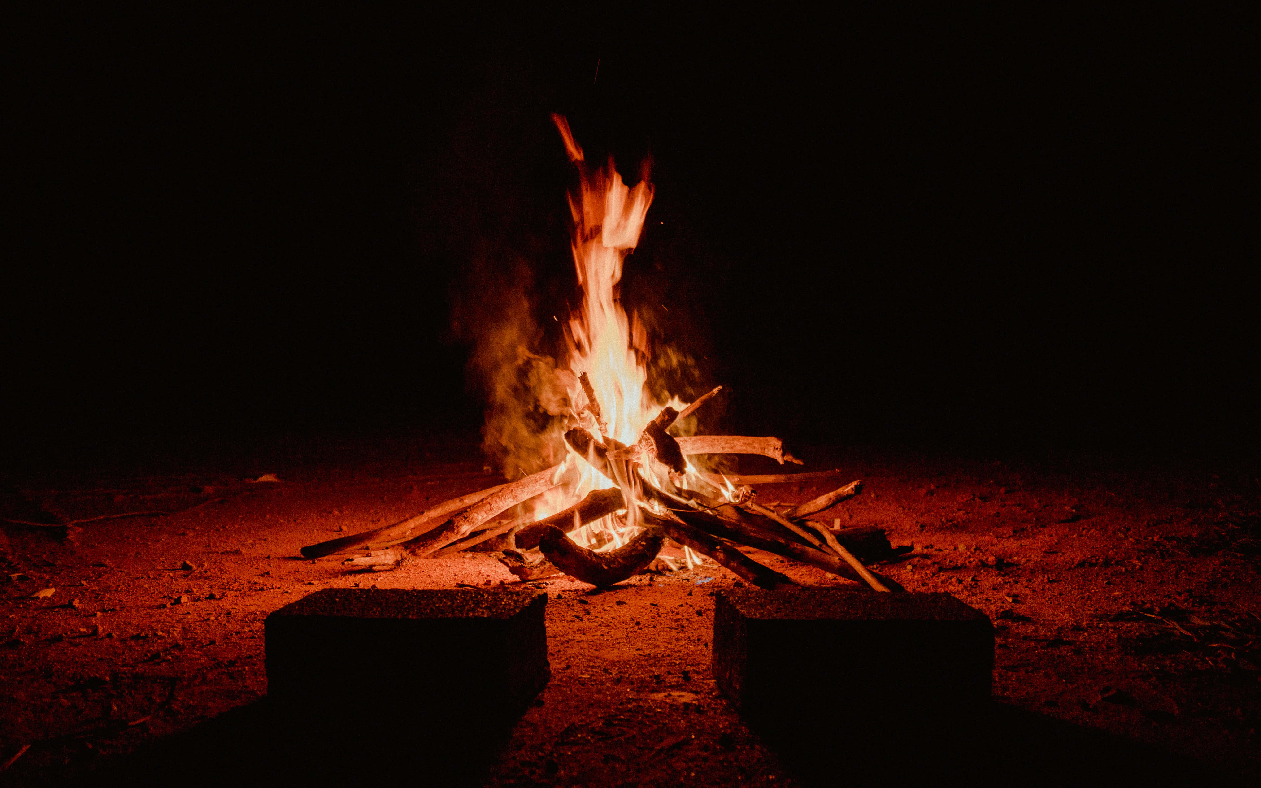 Bonfire 桌布 壁紙 — 熊熊營火在漆黑的夜空下，將溫暖的橙色光芒投射到周圍的泥土和兩塊石頭上。