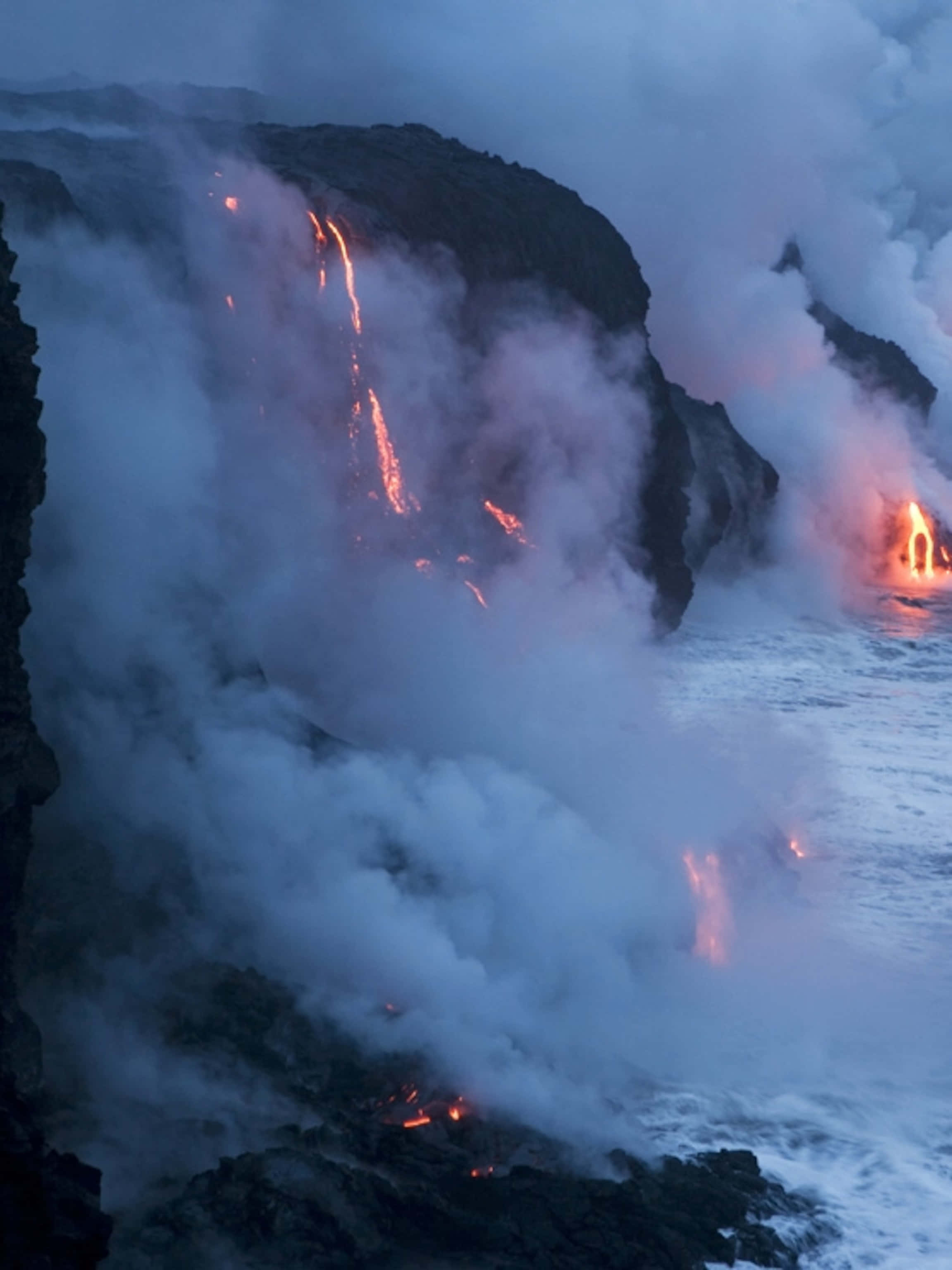 Volcano 桌布 壁紙 — 熔岩從懸崖傾瀉入海，在黑暗的暮色天空下產生蒸汽和發光的橙色痕跡。