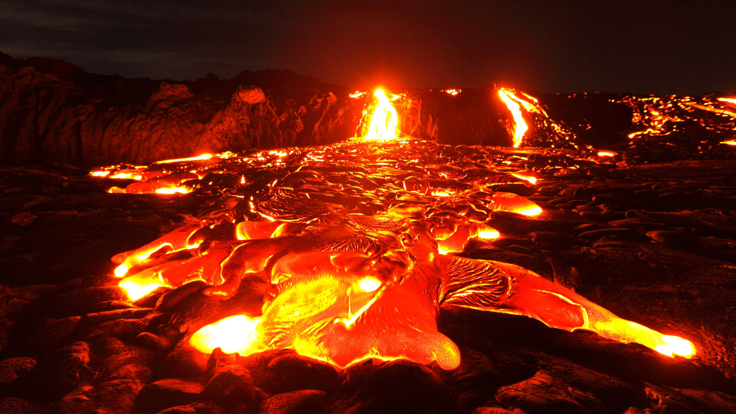 Volcano 桌布 壁紙 — 熔岩流在崎嶇地形上強烈發光，在黑暗的夜空下，遠處有熾熱的噴發。