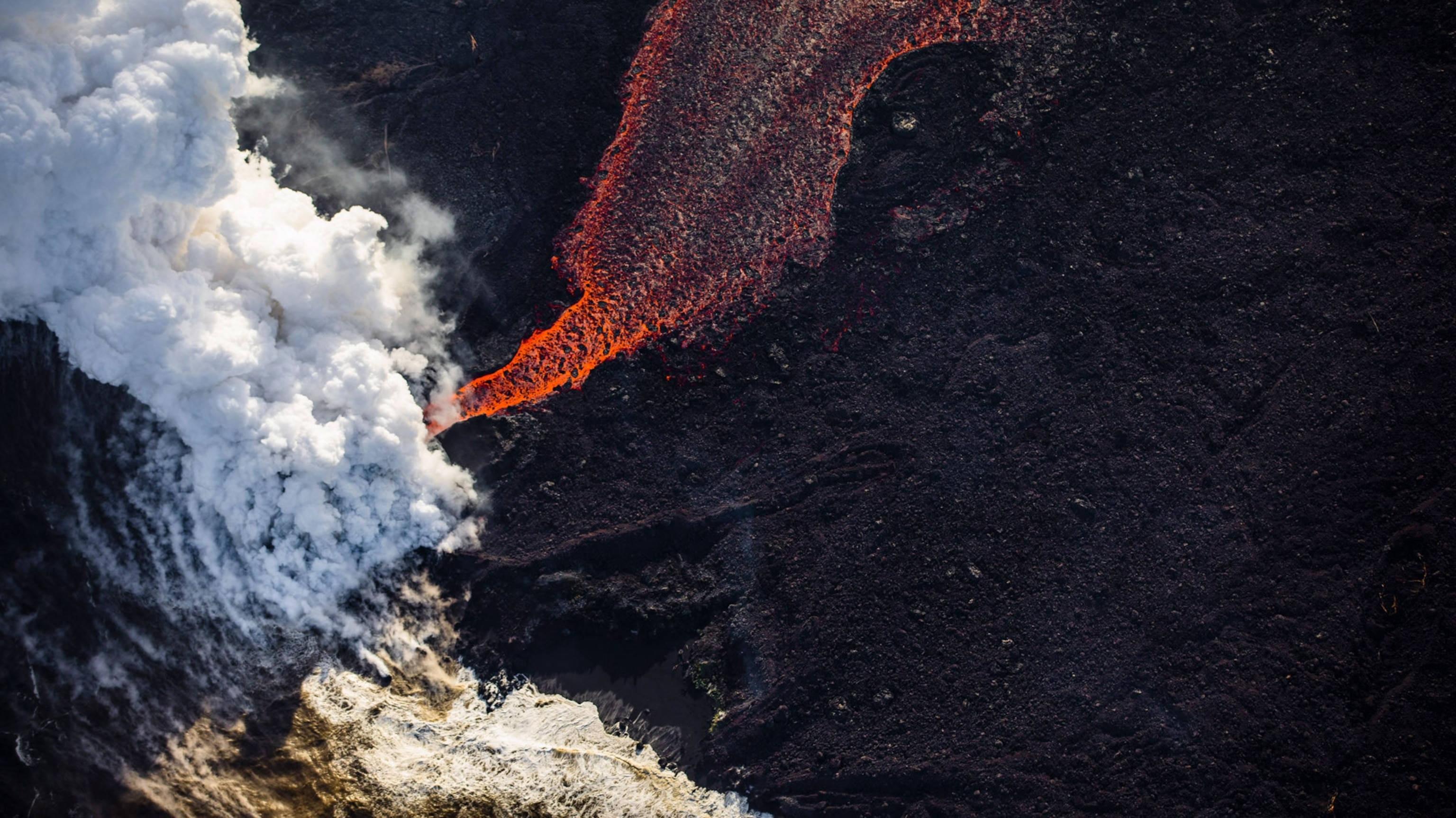 自然 桌布 壁紙 — 熔岩流過黑暗火山地形的鳥瞰圖，旁邊有滾滾白煙升起。