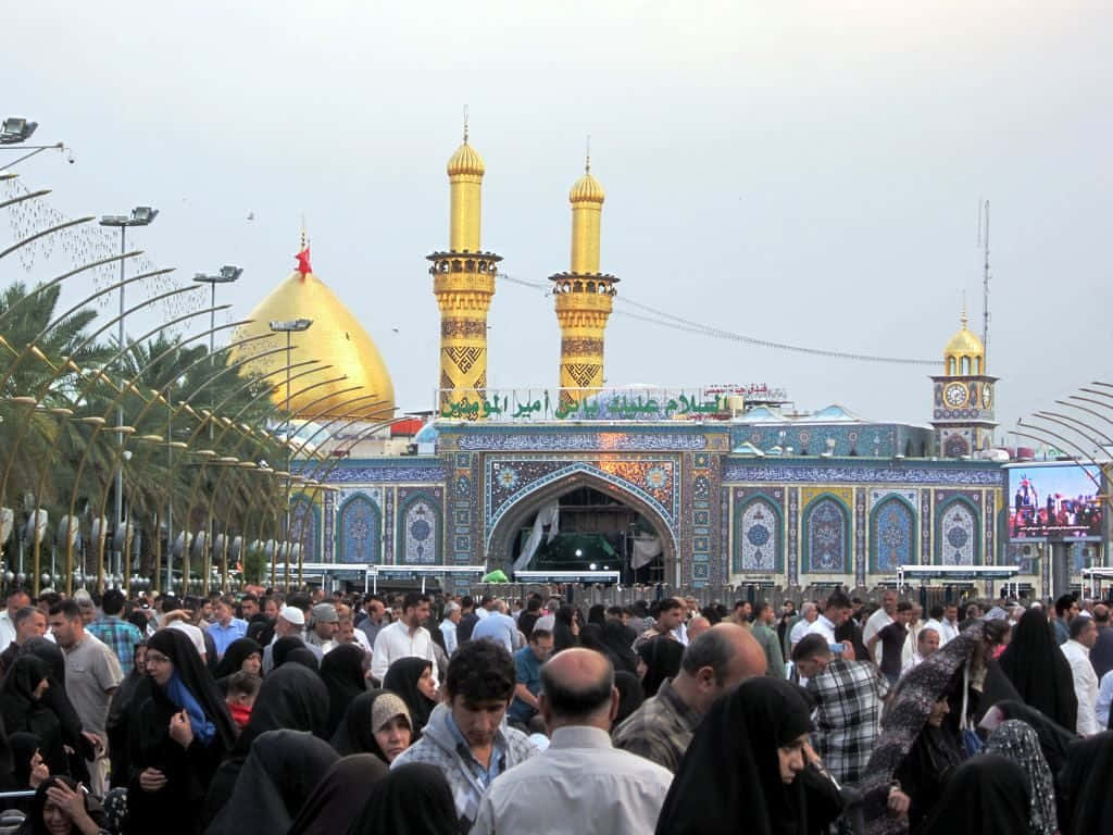 Karbala 桌布 壁紙 — 熙熙攘攘的人群聚集在一座宏偉的清真寺前，清真寺有金色圓頂和宣禮塔，天空多雲。