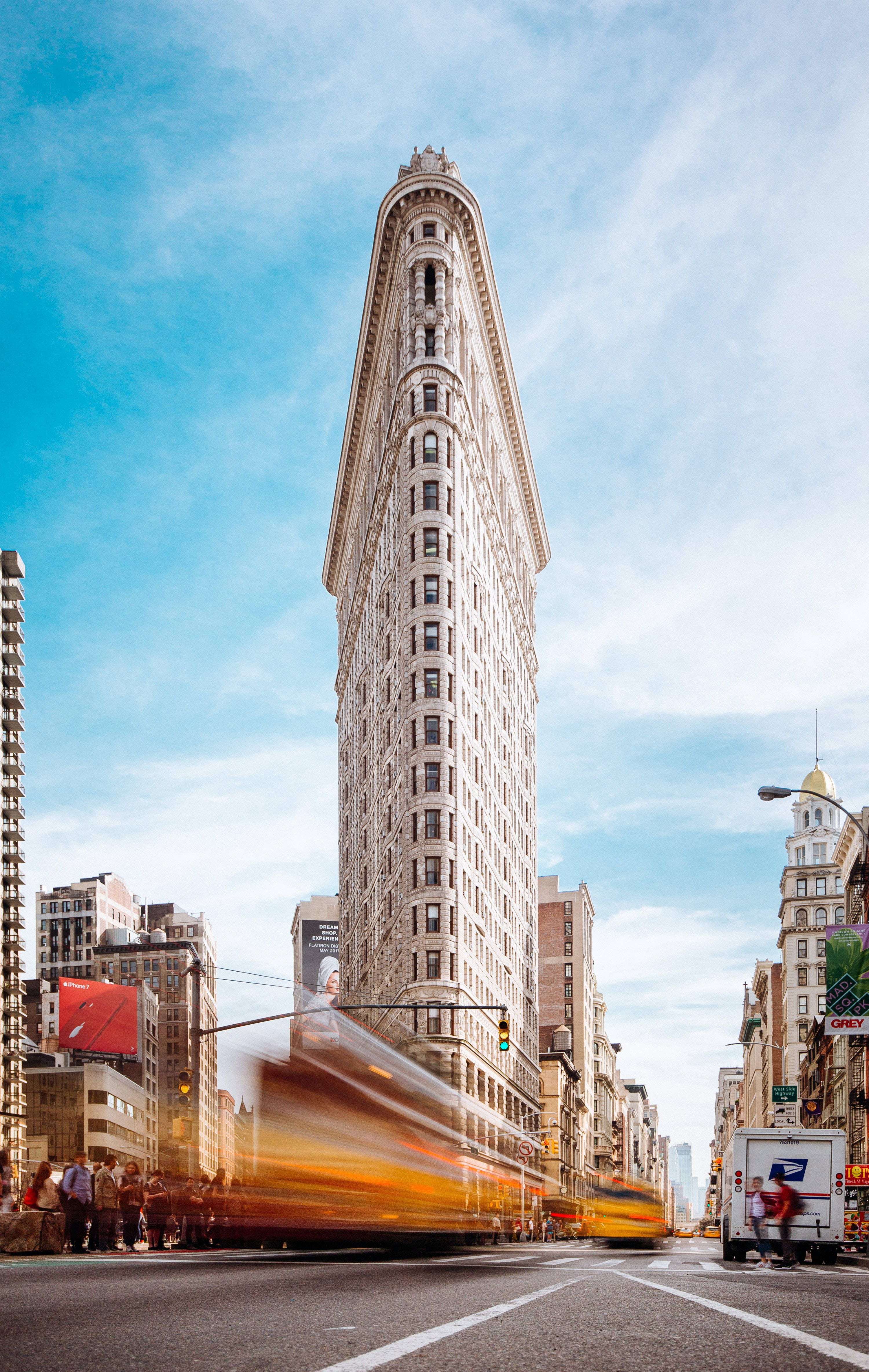 New York 桌布 壁紙 — 熨斗大廈在明亮的藍天下高聳矗立，一輛模糊的黃色巴士在繁忙的紐約街道上疾馳而過。