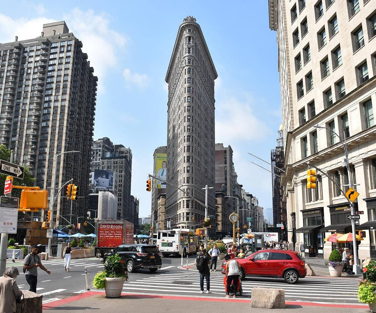 Flatiron Building 桌布 壁紙 — 熨斗大廈聳立在紐約市一條繁忙的街道上，街道上滿是行人、汽車和黃色交通燈，天空清澈湛藍。