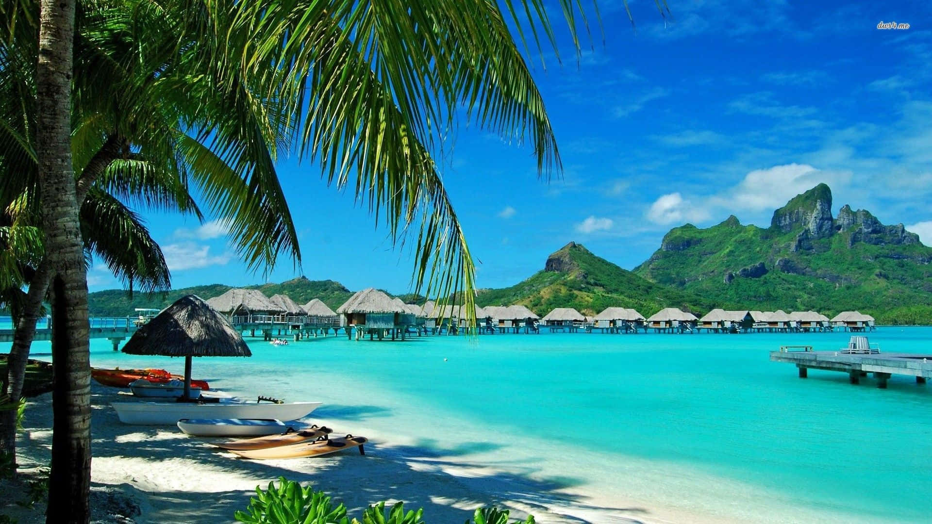 Hawaiian Beach 桌布 壁紙 — 熱帶天堂，擁有碧綠潟湖、棕櫚樹和水上平房，背景是鬱鬱蔥蔥的綠色山脈，天空湛藍明亮。
