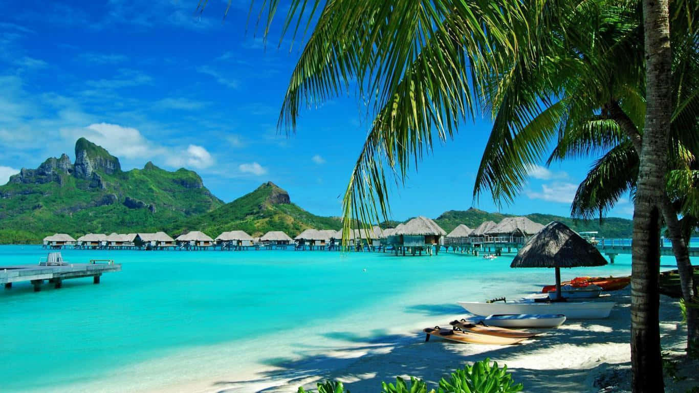 Hawaii Beach 桌布 壁紙 — 熱帶天堂，擁有碧綠色的潟湖、水上平房、棕櫚樹和鬱鬱蔥蔥的綠色山脈，在明亮的藍天下。