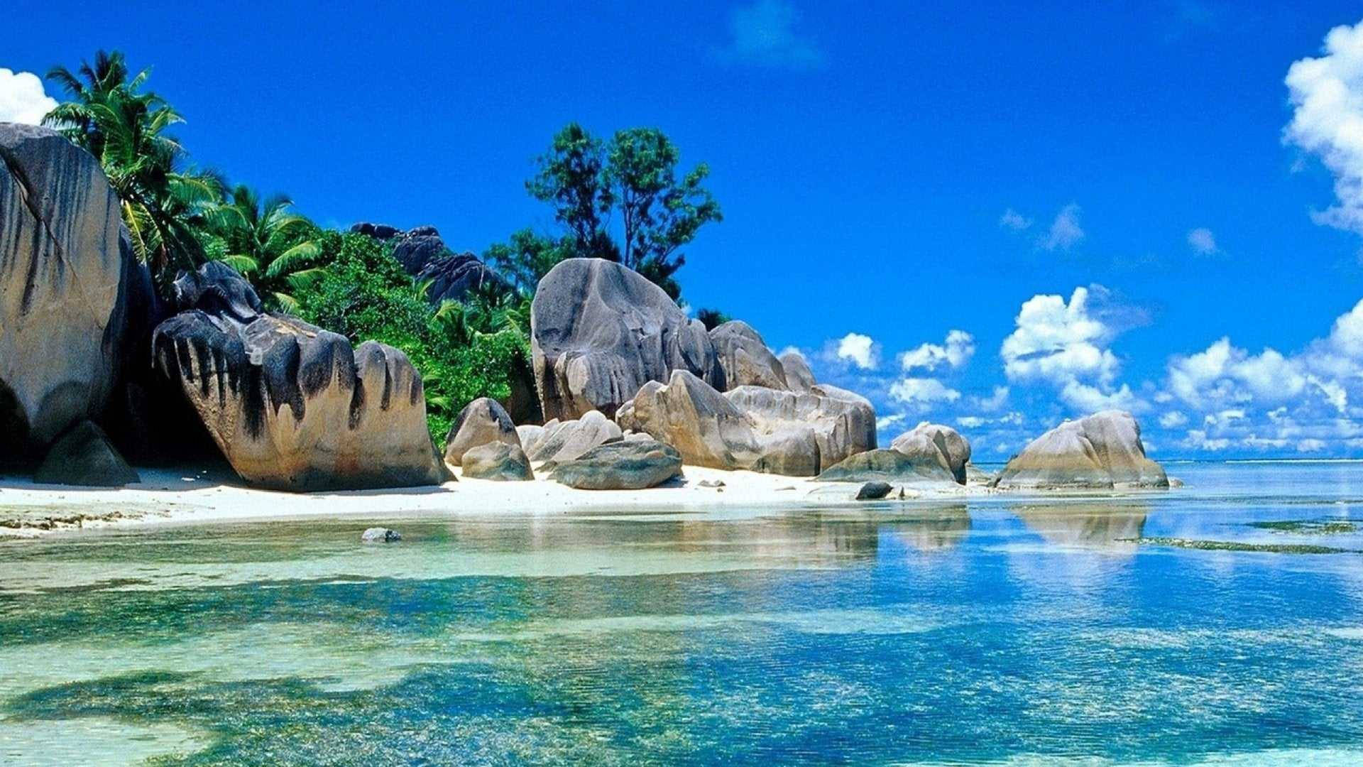 Best Summer 桌布 壁紙 — 熱帶天堂，碧綠的海水拍打著白色沙灘，周圍環繞著巨石和茂密的棕櫚樹，頭頂是明亮的藍天。