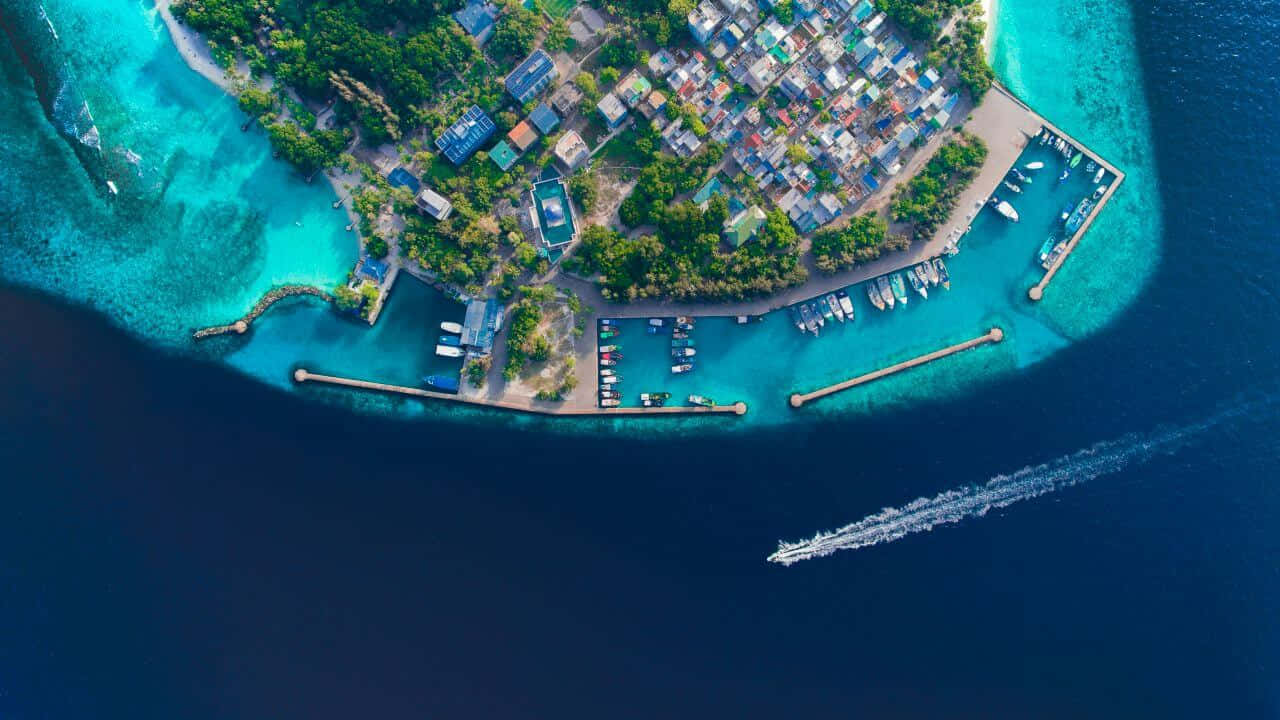 Maldives Island 桌布 壁紙 — 熱帶島嶼的鳥瞰圖，有藍綠色潟湖、船隻、碼頭和色彩繽紛的房屋，坐落在綠樹叢中。