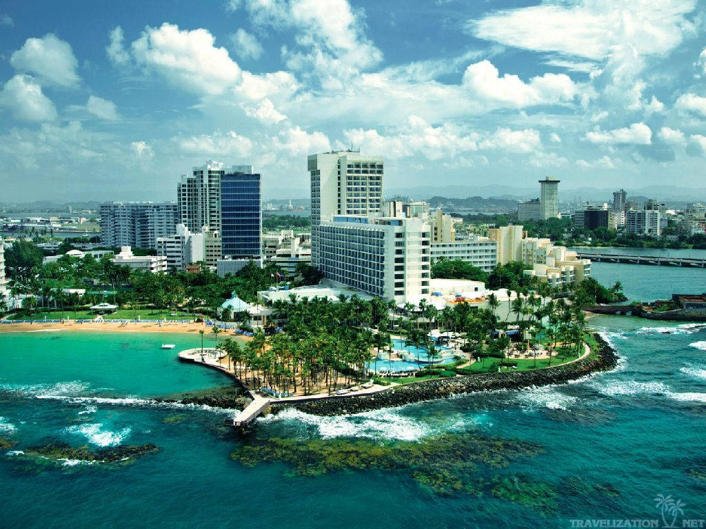 Puerto Rico 桌布 壁紙 — 熱帶沿海城市的鳥瞰圖，碧綠的海水拍打著岩石海岸，兩旁種滿棕櫚樹和現代酒店，明亮的藍天白雲。
