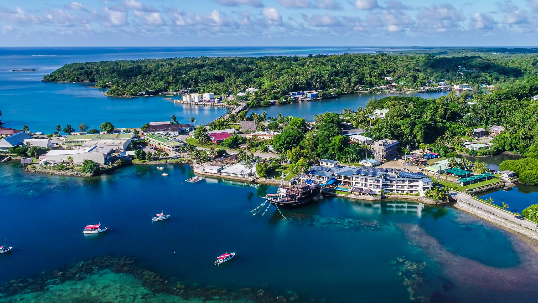 Micronesia 桌布 壁紙 — 熱帶沿海小鎮的鳥瞰圖，擁有碧綠海水、船隻和鬱鬱蔥蔥的綠色山丘，天空部分多雲。
