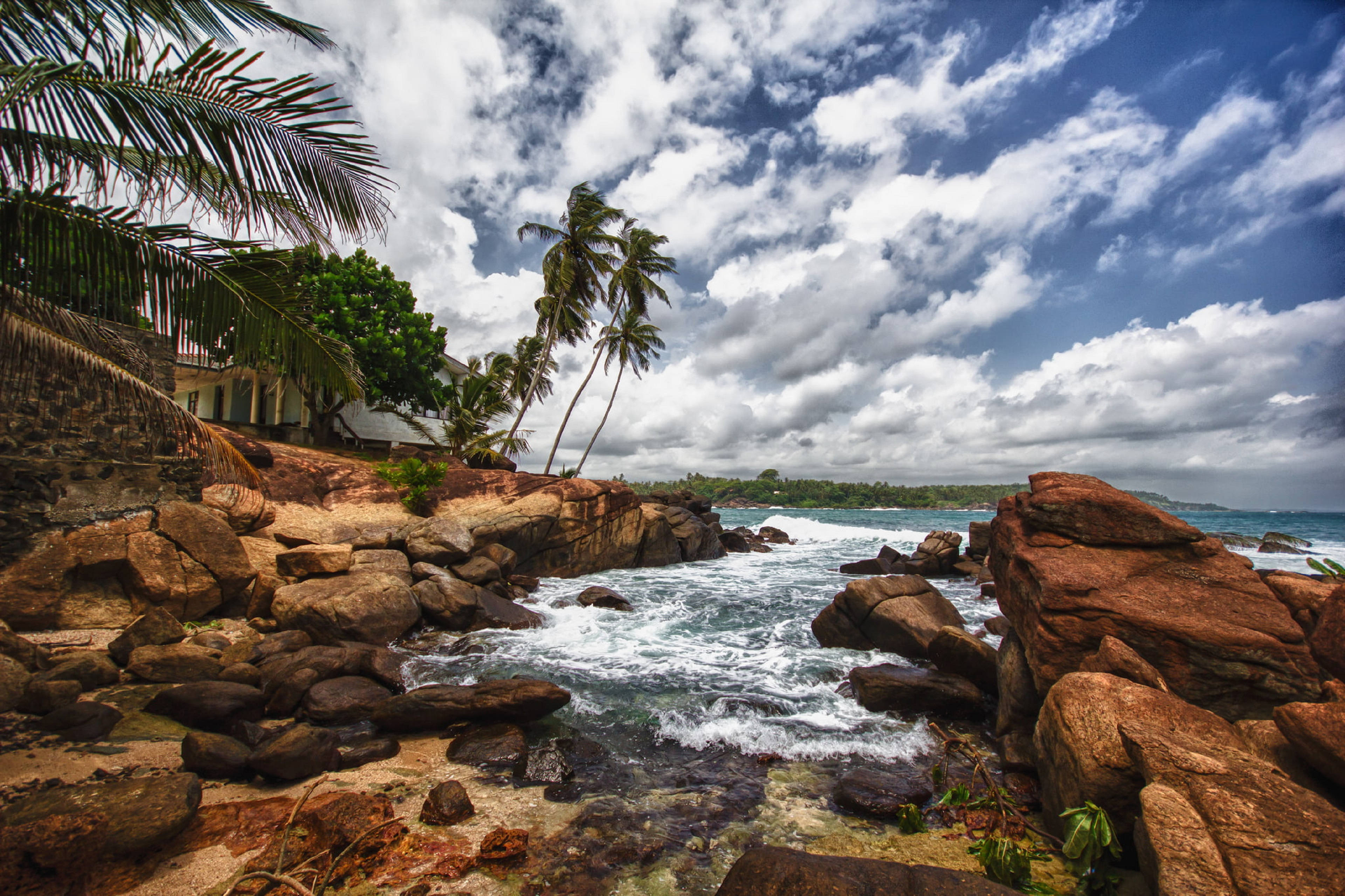 Sri Lanka 桌布 壁紙 — 熱帶海岸線，在戲劇性的多雲天空下，紅褐色岩石上拍打著浪花，棕櫚樹搖曳生姿。