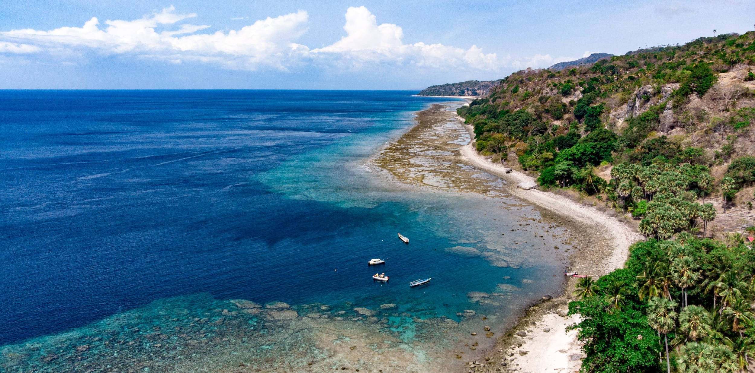 Timor Leste 桌布 壁紙 — 熱帶海岸線的鳥瞰圖，有碧綠的海水、珊瑚礁和鬱鬱蔥蔥的綠色山丘，在明亮的藍天下有蓬鬆的雲朵。