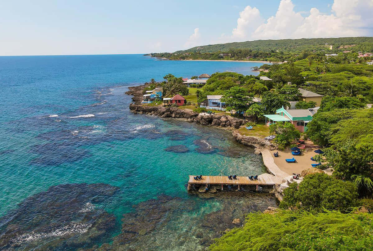 Jamaican Island 桌布 壁紙 — 熱帶海岸線的鳥瞰圖，有碧綠的海水、岩石海岸、茂密的綠色植物和色彩繽紛的海邊房屋，在明亮的藍天下。