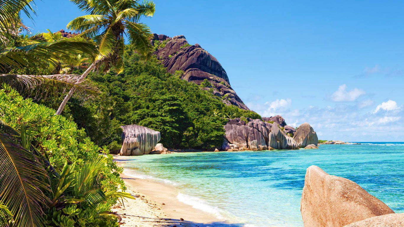 Seychelles 桌布 壁紙 — 熱帶海灘，有碧綠海水、沙灘、茂密的棕櫚樹和岩石懸崖，在明亮的藍天下。