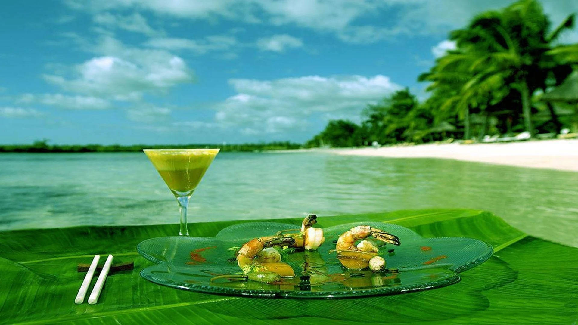Lunch 桌布 壁紙 — 熱帶海灘場景，一盤蝦和雞尾酒放在巨型綠葉上，俯瞰碧綠海水和棕櫚樹。