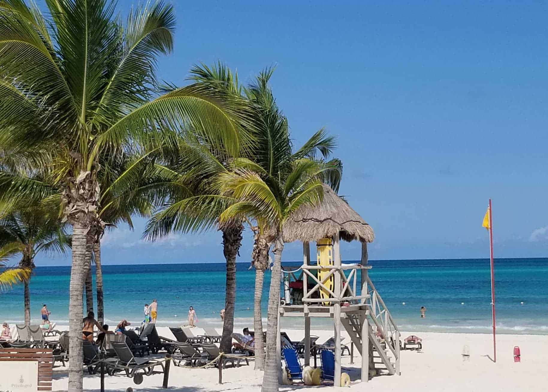 Mexican Beach 桌布 壁紙 — 熱帶海灘場景，棕櫚樹、躺椅和茅草救生塔在明亮的藍天下。