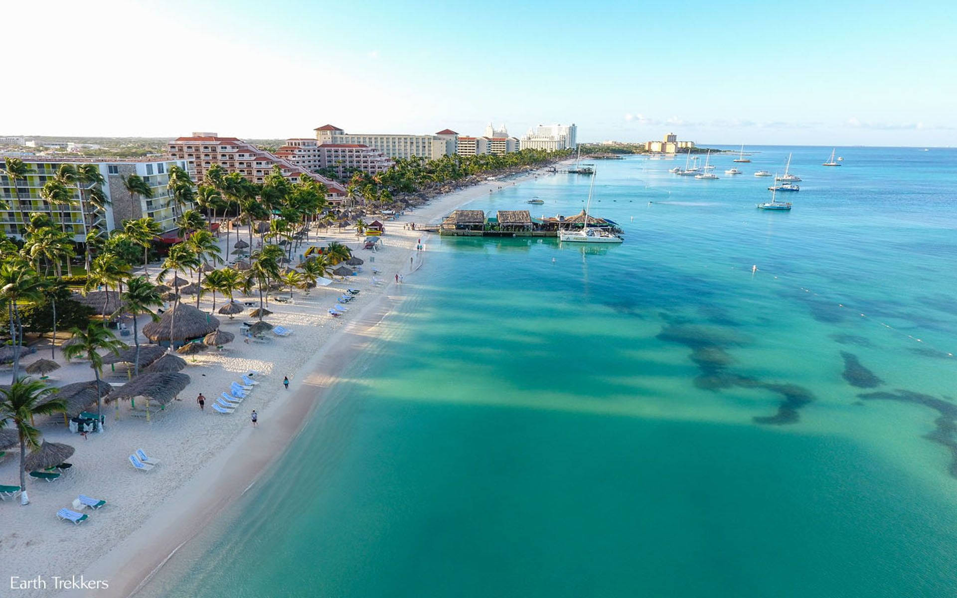 Aruba 桌布 壁紙 — 熱帶海灘的鳥瞰圖，有碧綠的海水、棕櫚樹、度假村建築和帆船，天空晴朗湛藍。