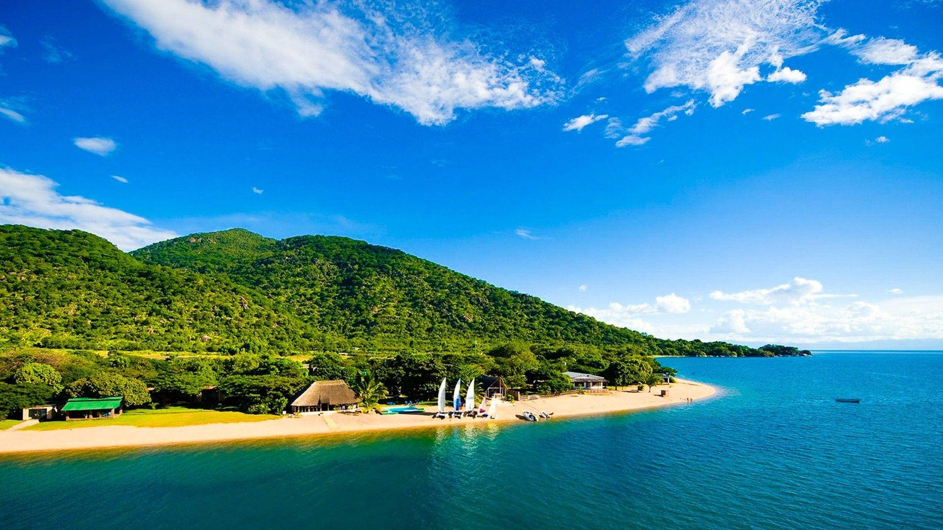 Mozambique 桌布 壁紙 — 熱帶海灘的鳥瞰圖，有碧綠的海水、綠色的山丘和白色的帆船，在明亮的藍天下。