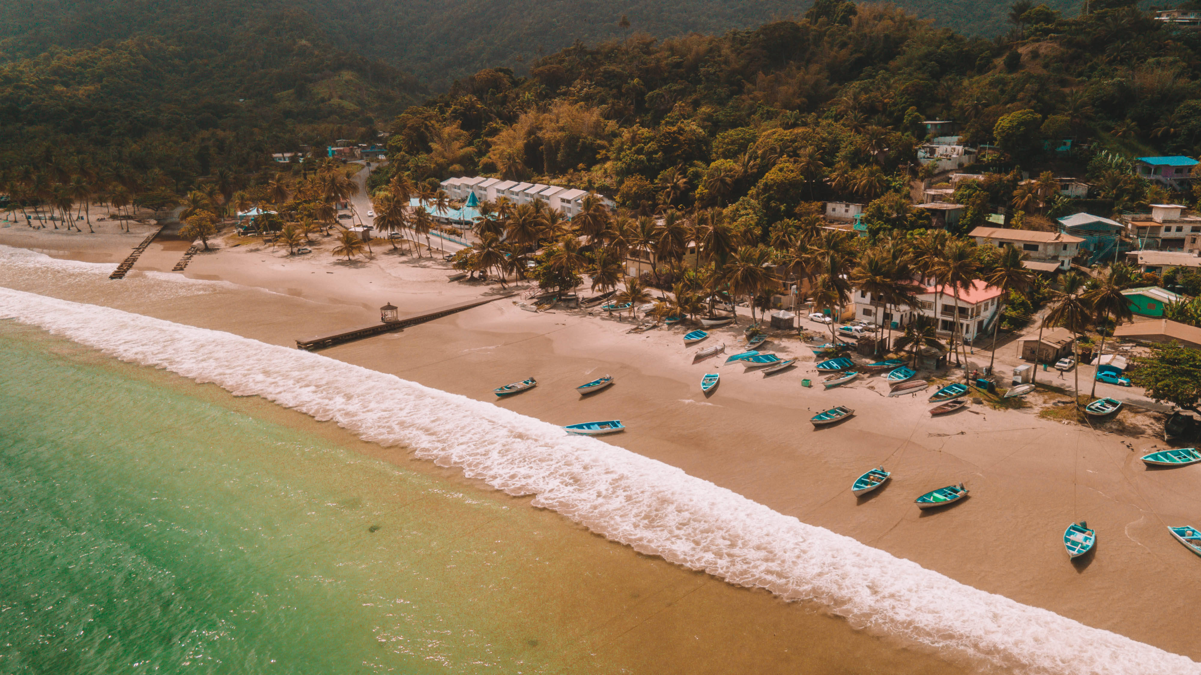 Trinidad 桌布 壁紙 — 熱帶海灘的鳥瞰圖，碧綠的海浪拍打著沙灘，沙灘上點綴著棕櫚樹和散落的藍色船隻。