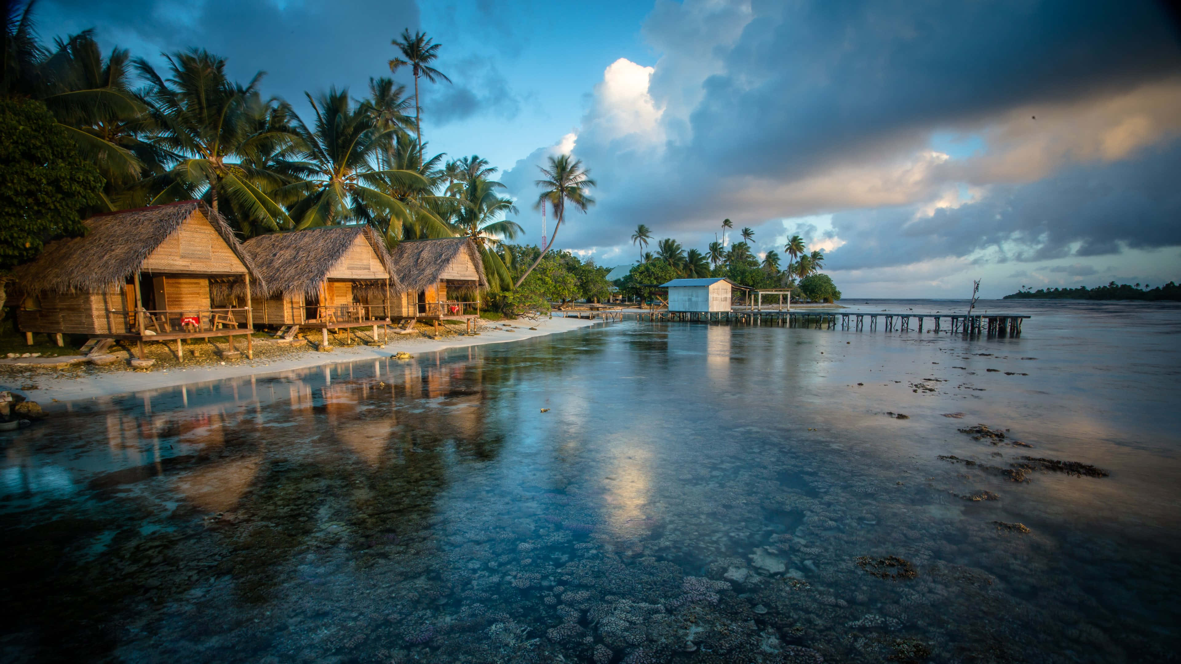 Resort 桌布 壁紙 — 熱帶海灘茅草屋頂小屋在戲劇性的暮色天空下倒映在平靜的碧綠海水中。
