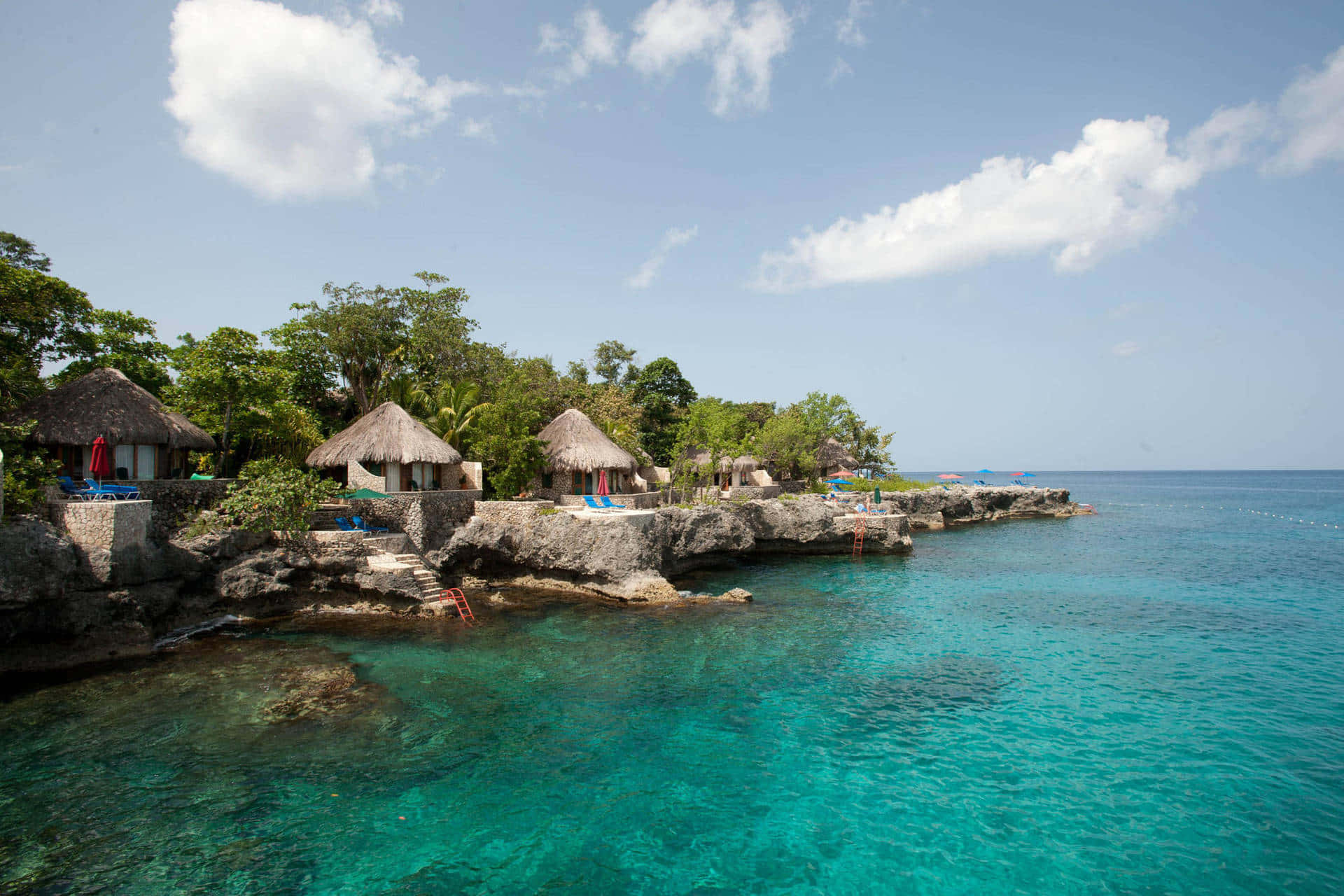 Jamaica 桌布 壁紙 — 熱帶茅草小屋坐落在岩石懸崖上，俯瞰著清澈碧綠的海水，頭頂是明亮的藍天和蓬鬆的白雲。