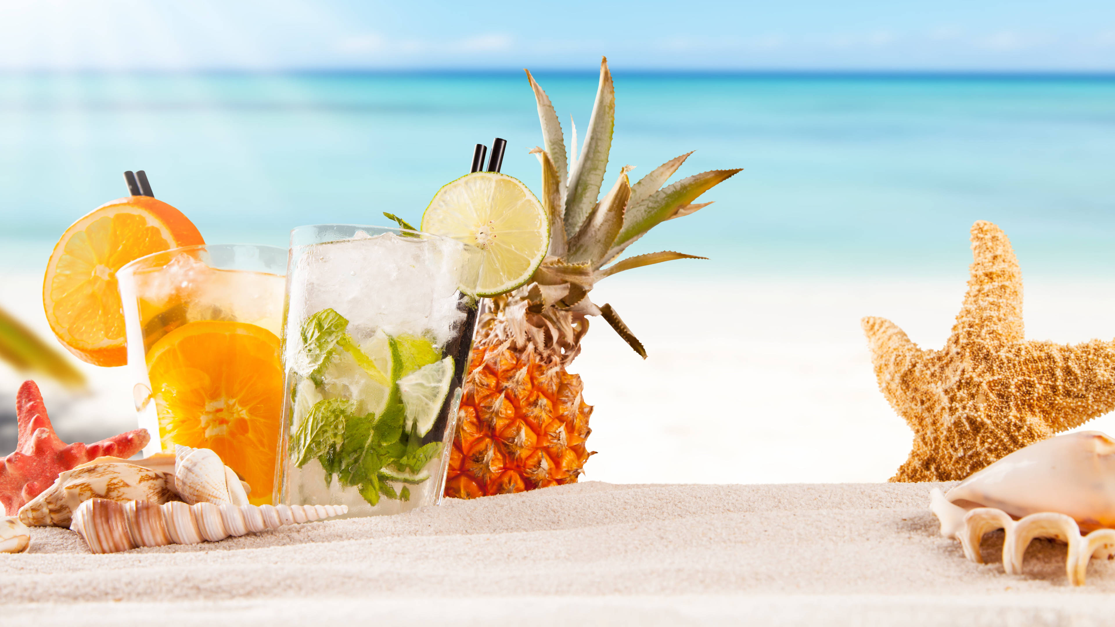 Tropical Drink 桌布 壁紙 — 熱帶雞尾酒配以鳳梨、柳橙和薄荷裝飾，放在沙灘上，旁邊有海星和貝殼，在明亮的藍天下。