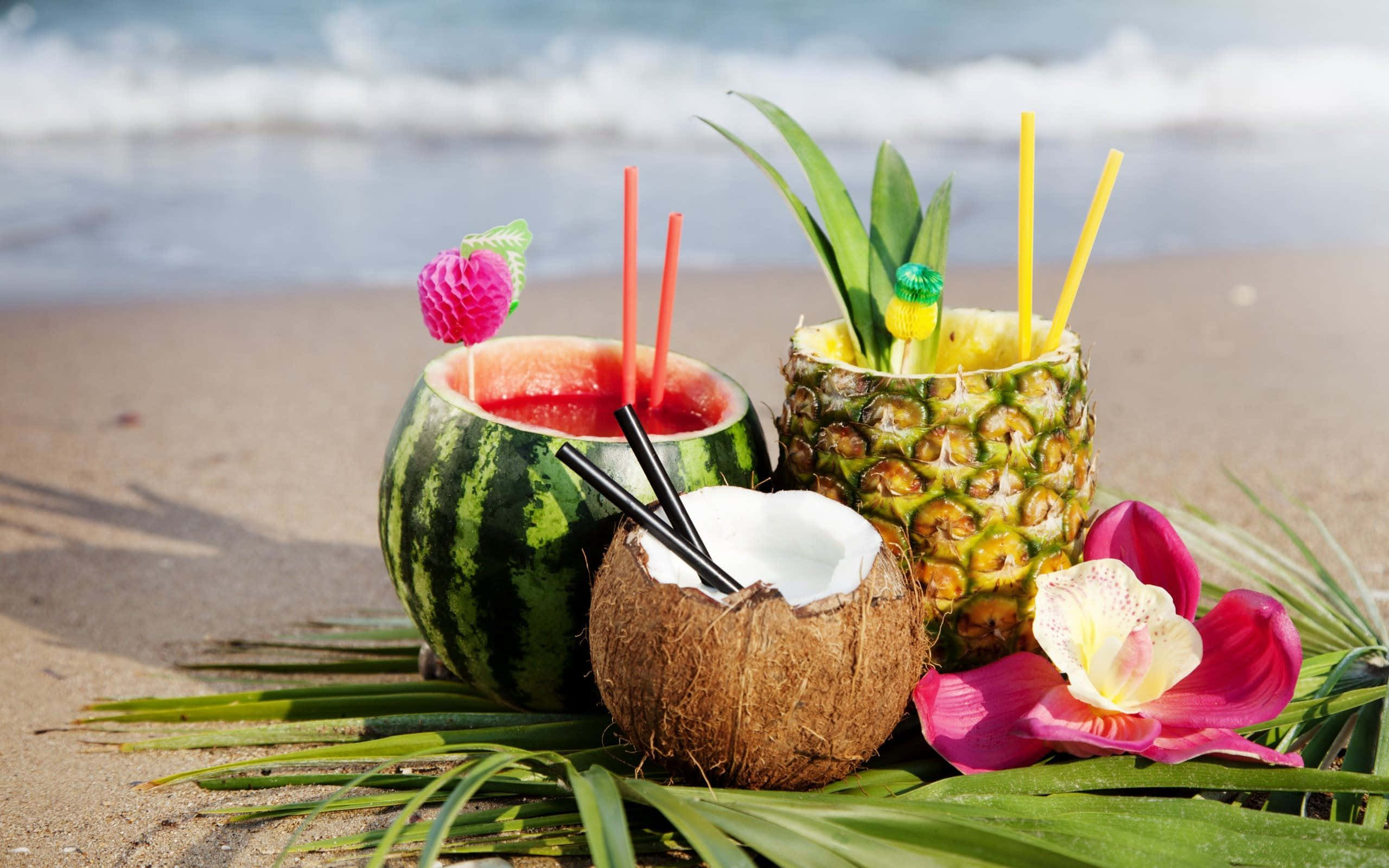 Coconut 桌布 壁紙 — 熱帶飲品放在西瓜、鳳梨和椰子殼中，置於沙灘上，背後有海浪。