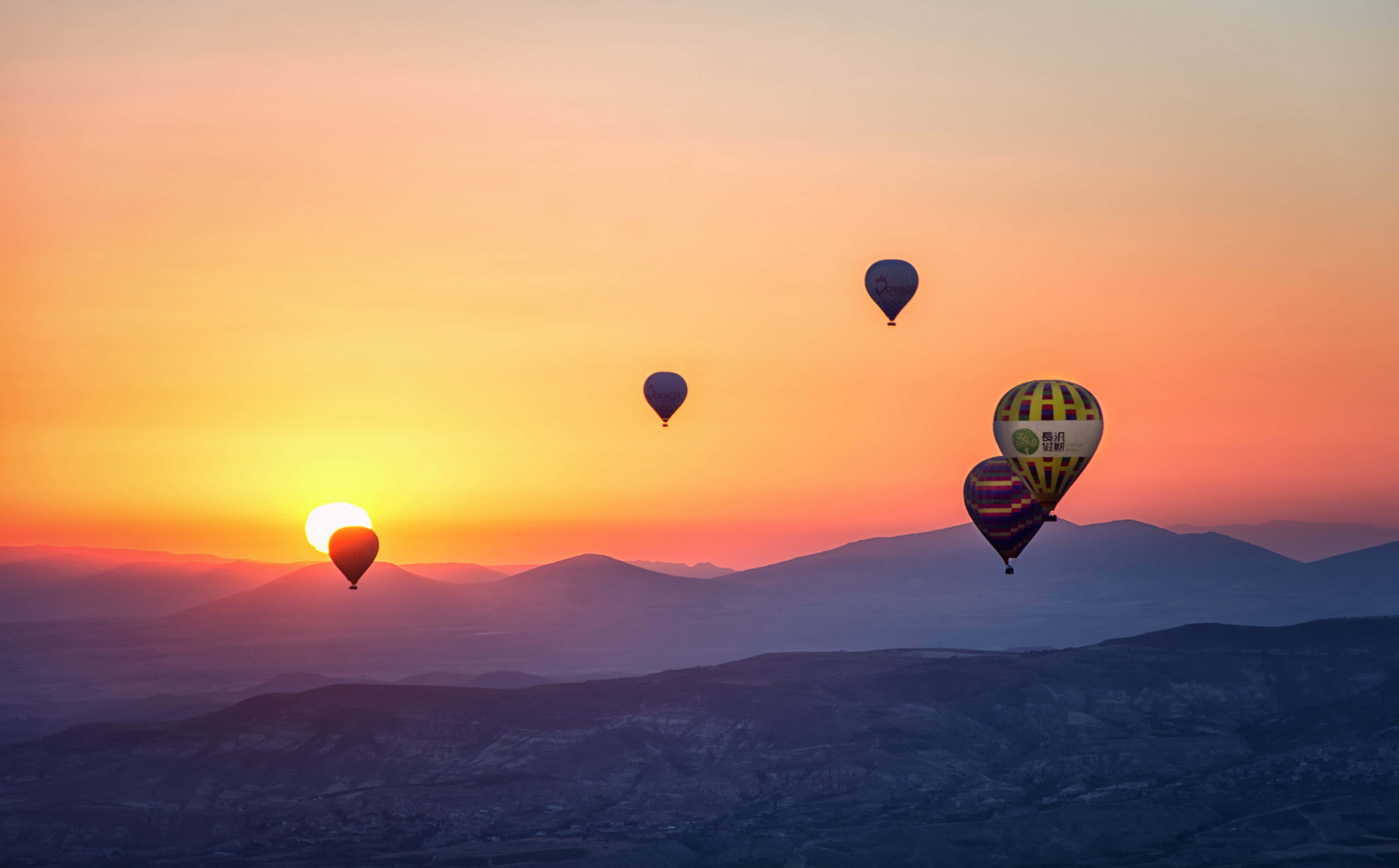 Hot Air Balloon 桌布 壁紙 — 熱氣球在山脈上空飄移，夕陽以火熱的橙色和粉紅色調落下。