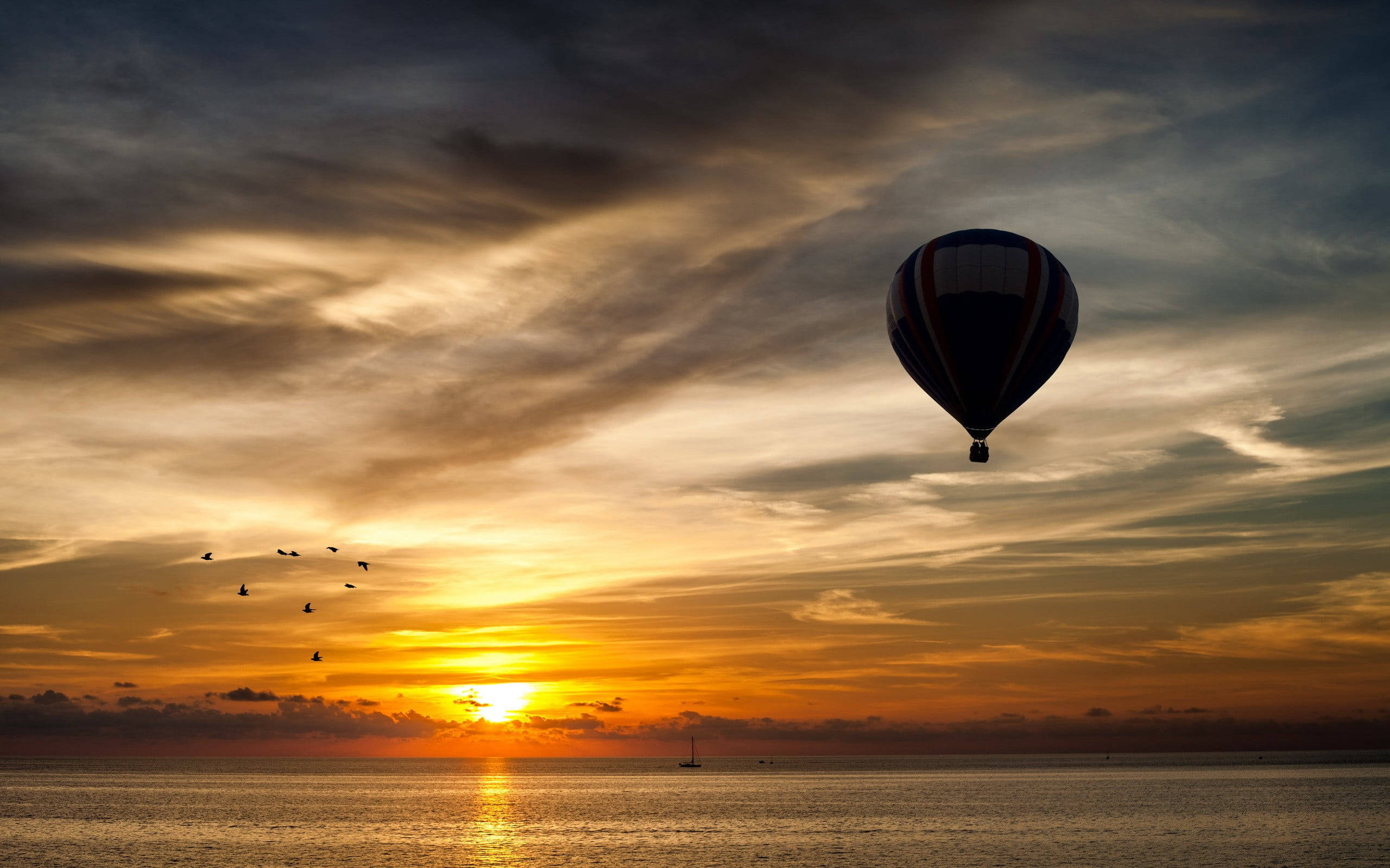 Hot Air Balloon 桌布 壁紙 — 熱氣球在平靜的海面上以熾熱的日落為背景形成剪影，鳥兒成群結隊飛翔。