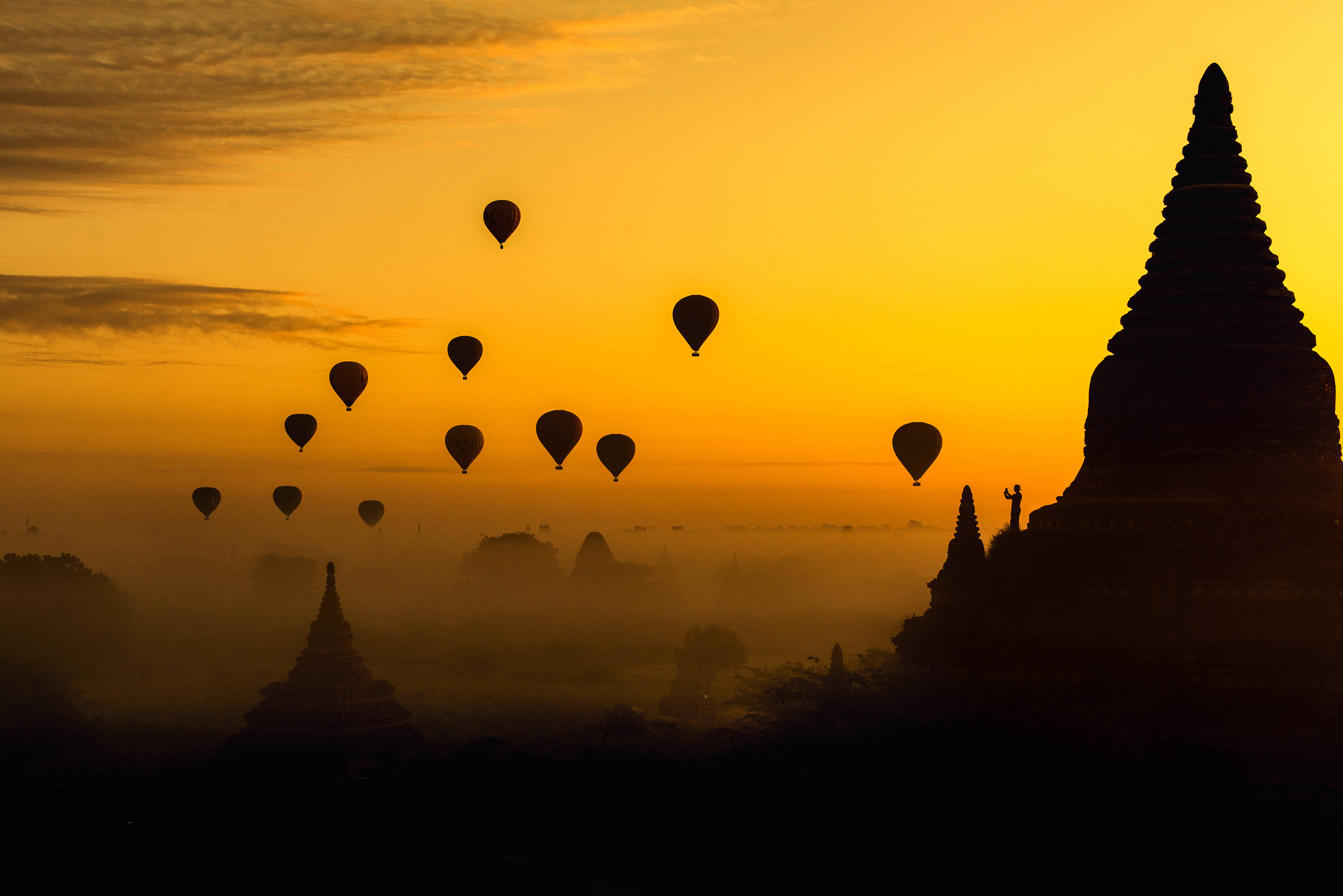 Burma 桌布 壁紙 — 熱氣球在霧氣籠罩的古老寶塔上空飄浮，金色日出在溫暖的天空中投射出戲劇性的剪影。