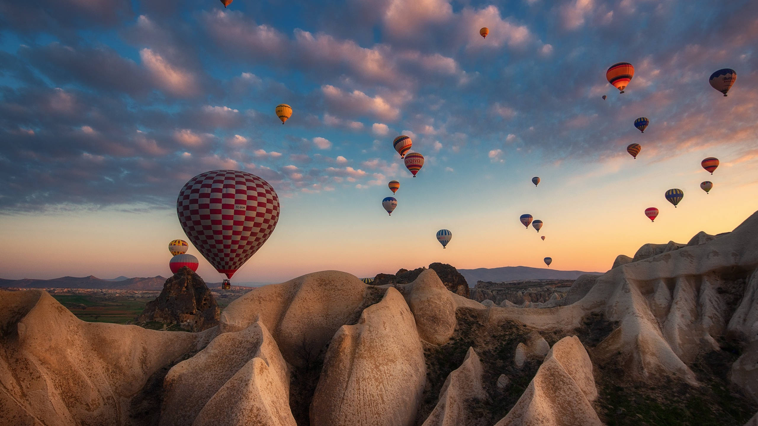 Hot Air Balloon 桌布 壁紙 — 熱氣球在黎明時分飄過超現實的岩石地貌，天空呈現柔和的粉紅色和橙色。