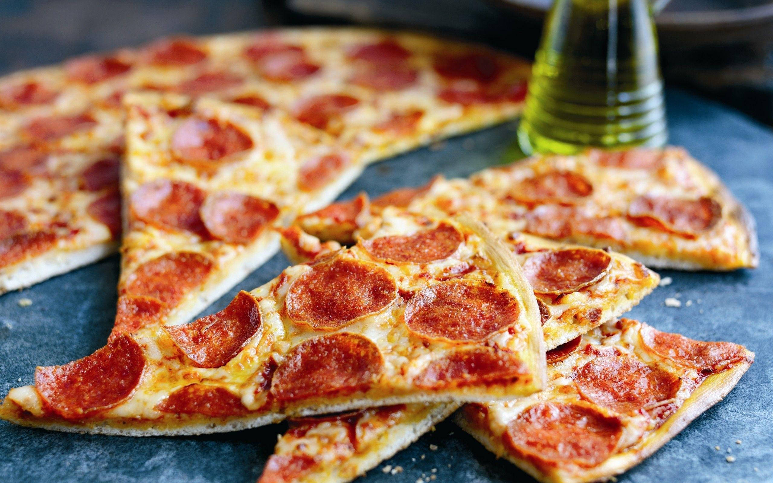 Pizza 桌布 壁紙 — 熱騰騰的義大利辣香腸披薩片高高堆在深色石板表面上，旁邊有一個綠色橄欖油瓶。