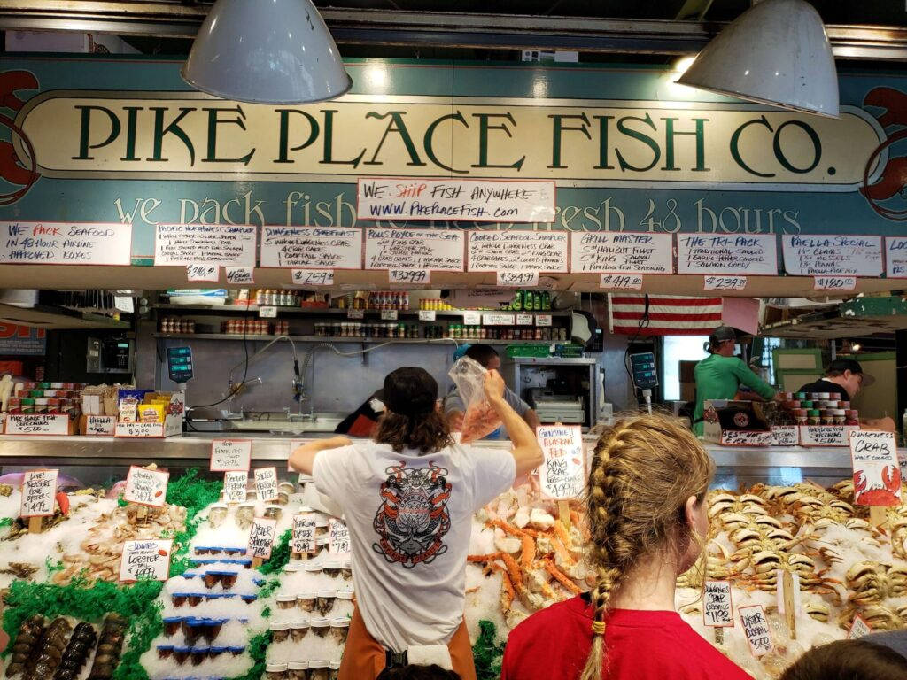 Pike Place Market 桌布 壁紙 — 熱鬧的 Pike Place Fish Co. 攤位，顧客和員工在明亮的燈光下處理新鮮海鮮。