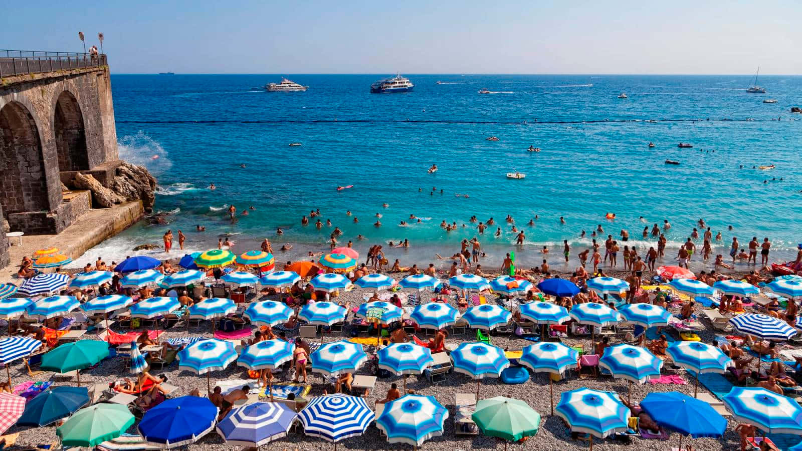 Italian Beach 桌布 壁紙 — 熱鬧的卵石灘，擠滿色彩繽紛的遮陽傘和享受碧綠海水的曬太陽人士，天空晴朗蔚藍。