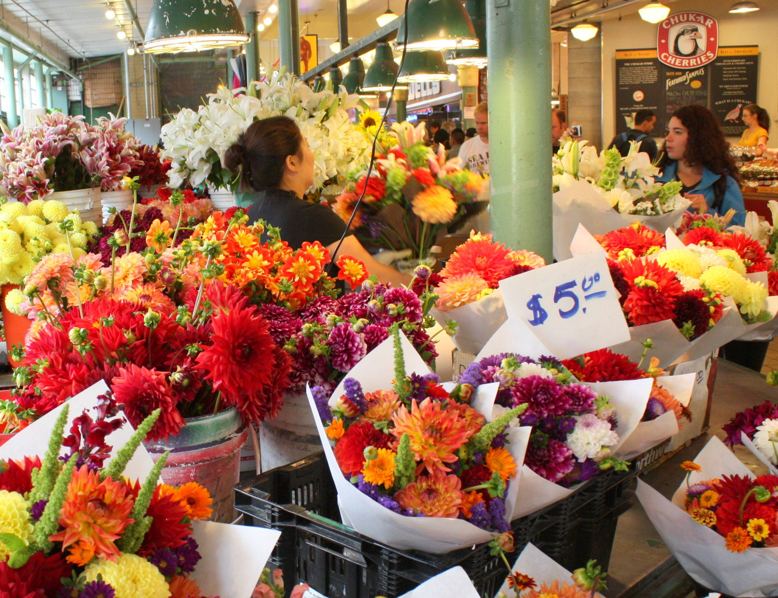 Pike Place Market 桌布 壁紙 — 熱鬧的室內市場中，色彩繽紛的花攤堆滿了紅色、橙色和紫色的大麗花。