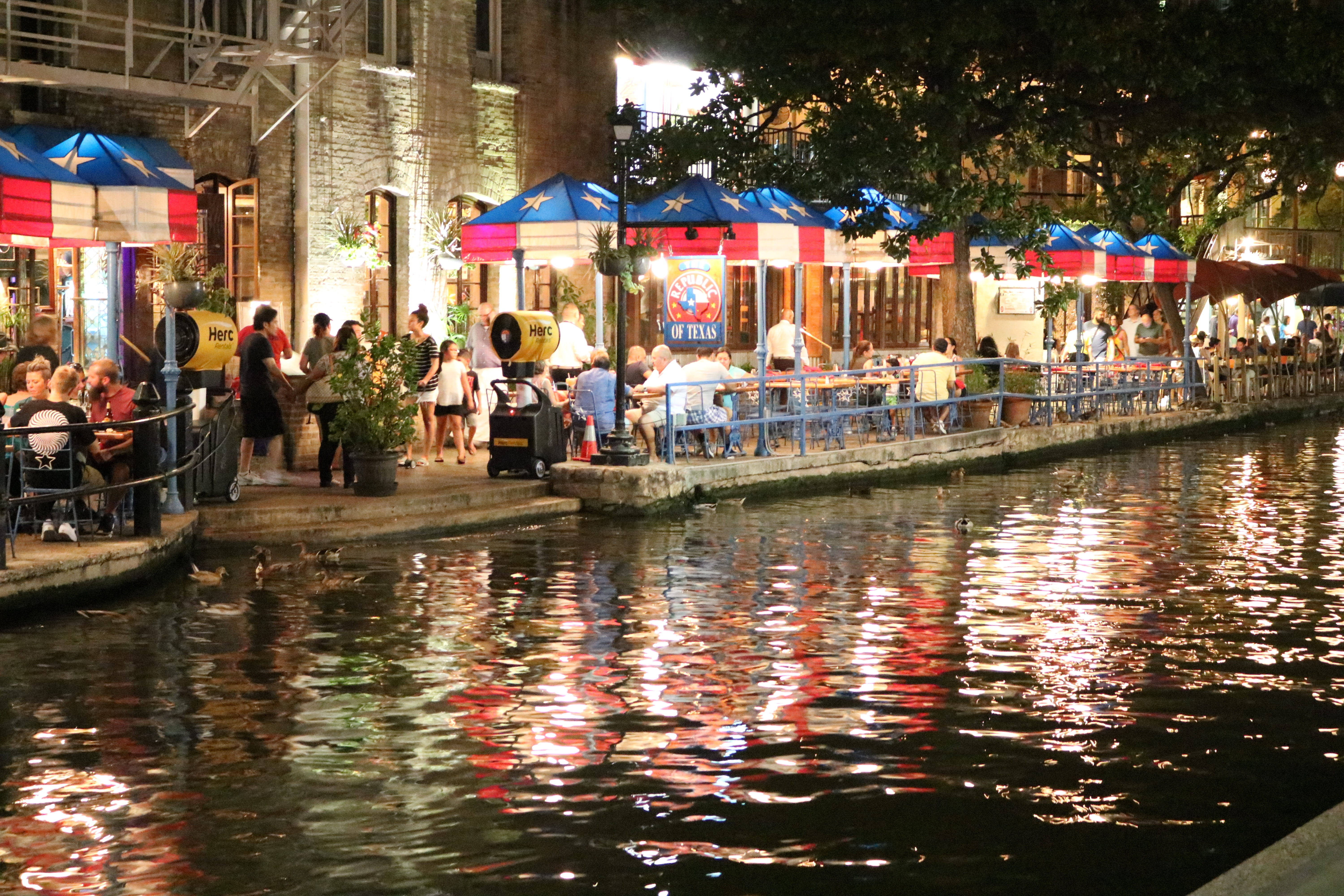 水 桌布 壁紙 — 熱鬧運河的夜景，人們在德州旗幟遮陽傘下用餐，五彩燈光倒映在水面上。