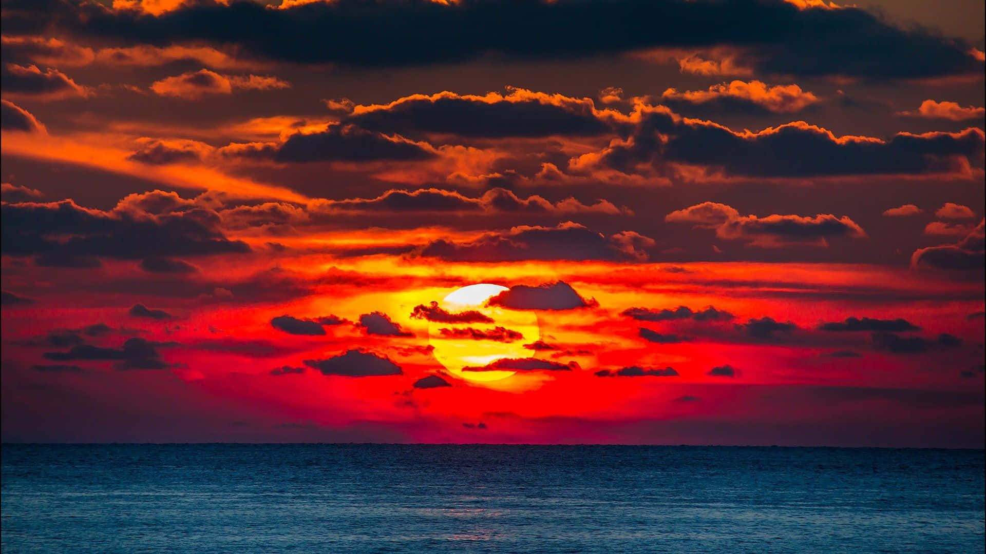 Orange Sunset 桌布 壁紙 — 熾熱的夕陽在平靜的海洋上空，將天空染成鮮豔的紅色和橙色，太陽沉入地平線之下。