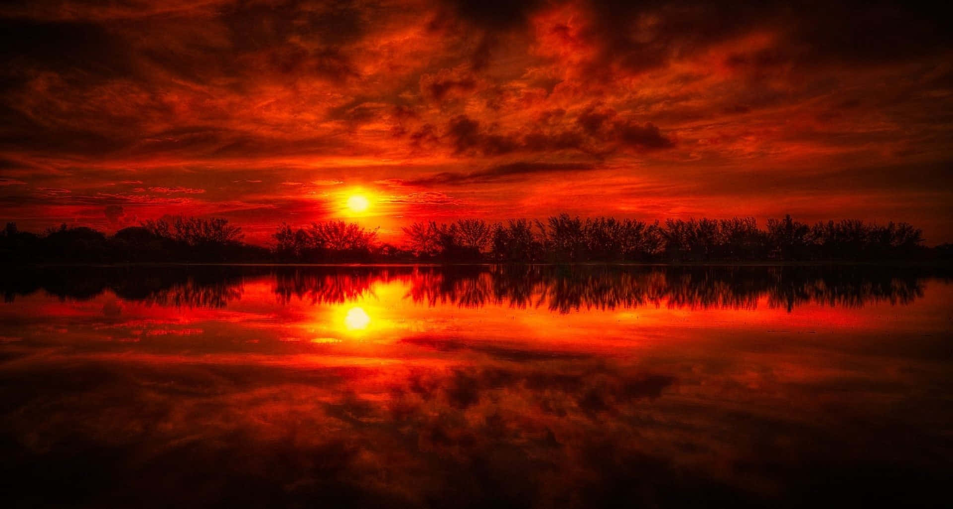 Red Sunset 桌布 壁紙 — 熾熱的夕陽將天空和水面染成鮮豔的紅色和橙色，地平線上有樹木的剪影。