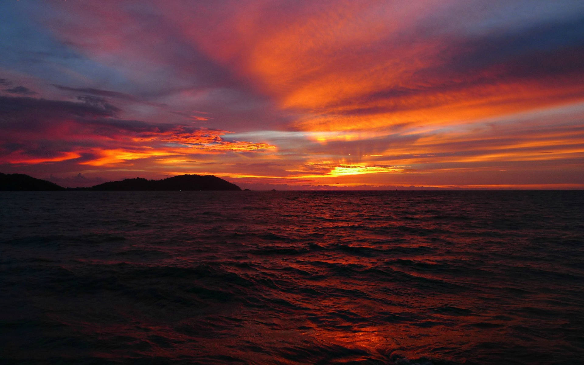Ocean Sunset 桌布 壁紙 — 熾熱的夕陽將天空和海洋染成鮮豔的紅色、橙色和紫色，雲彩在剪影般的島嶼上方劃過。