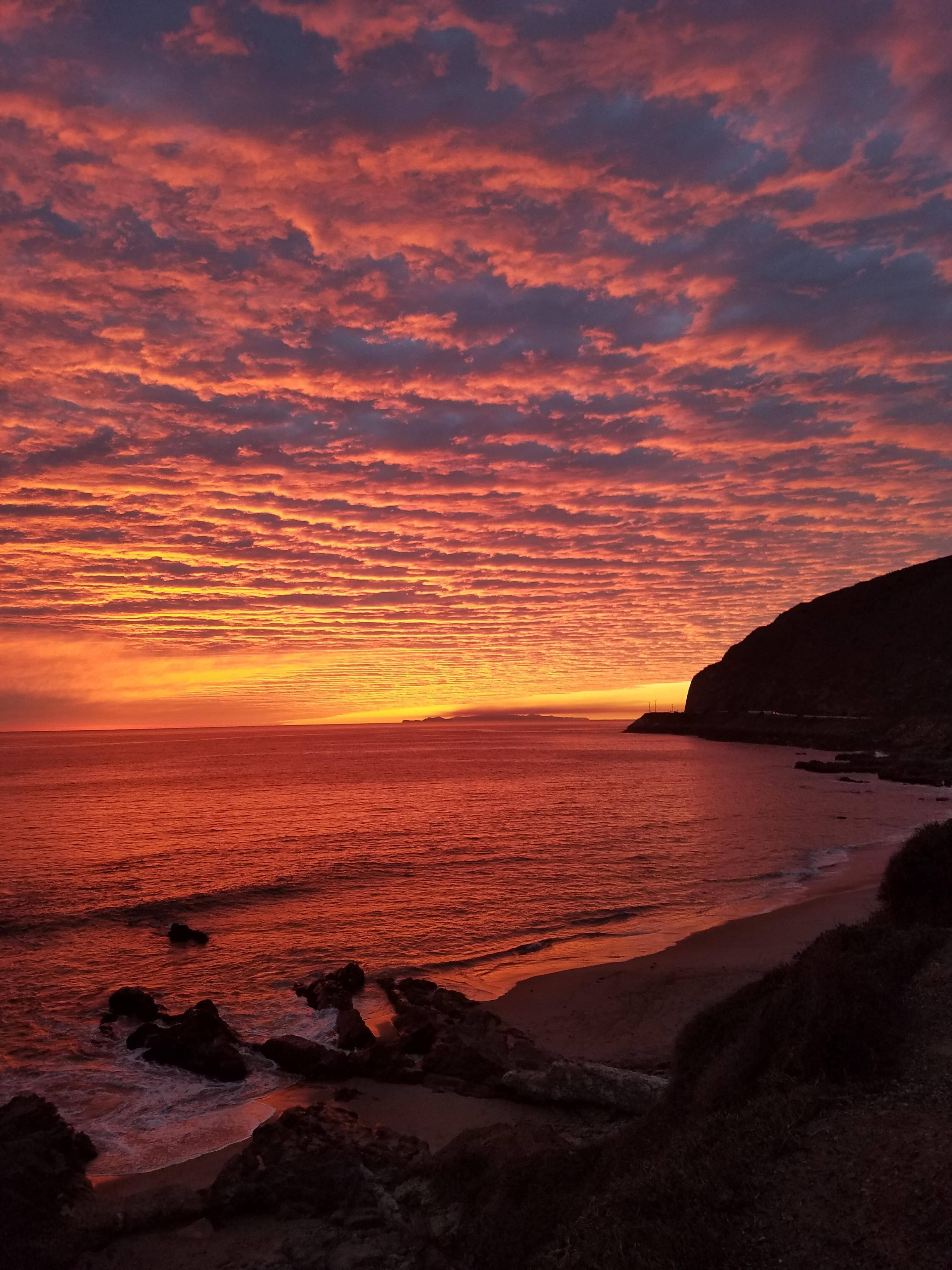 malibu iPhone 桌布 壁紙 — 熾熱的夕陽將天空和海洋染成鮮豔的紅色、橙色和紫色，海浪拍打著岩石海岸。