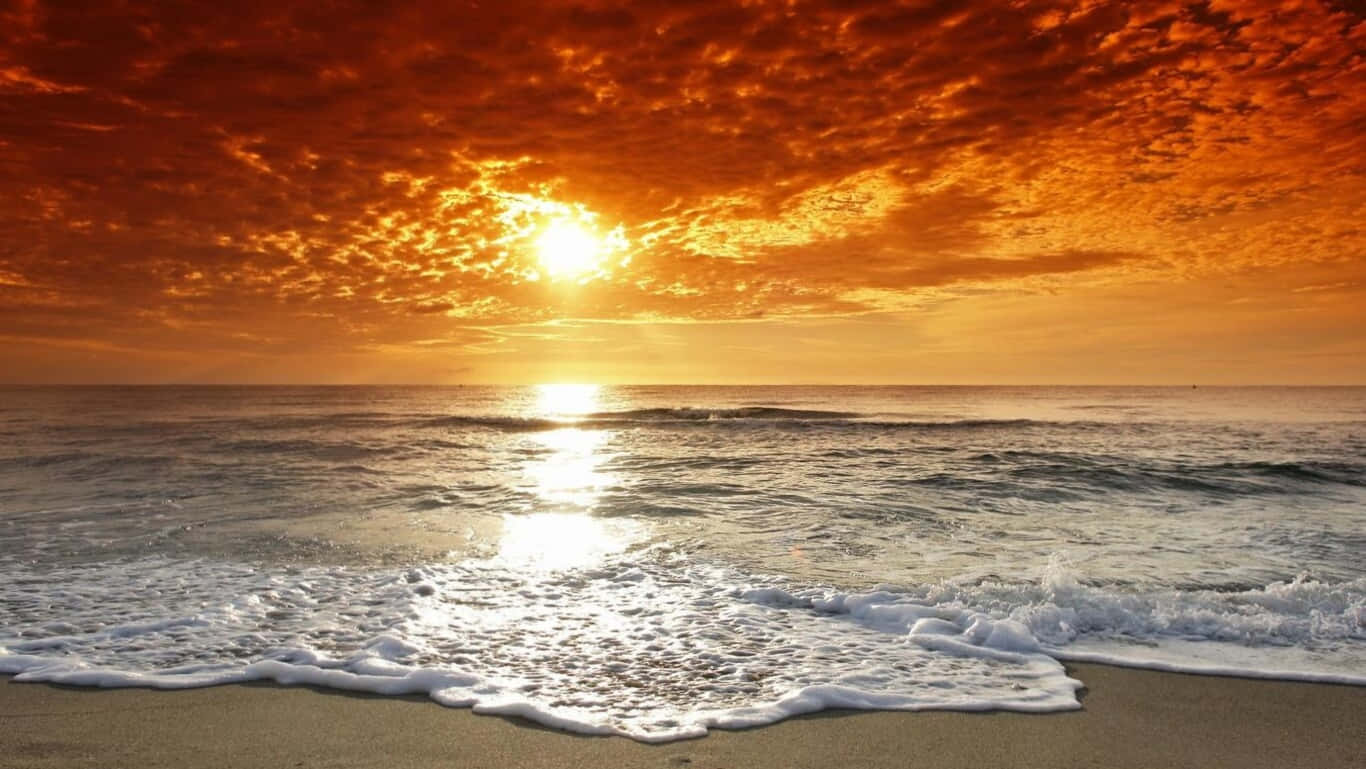 HD Beach 桌布 壁紙 — 熾熱的夕陽將天空染成橙色，海浪拍打沙灘，金色的光芒在水面上反射。