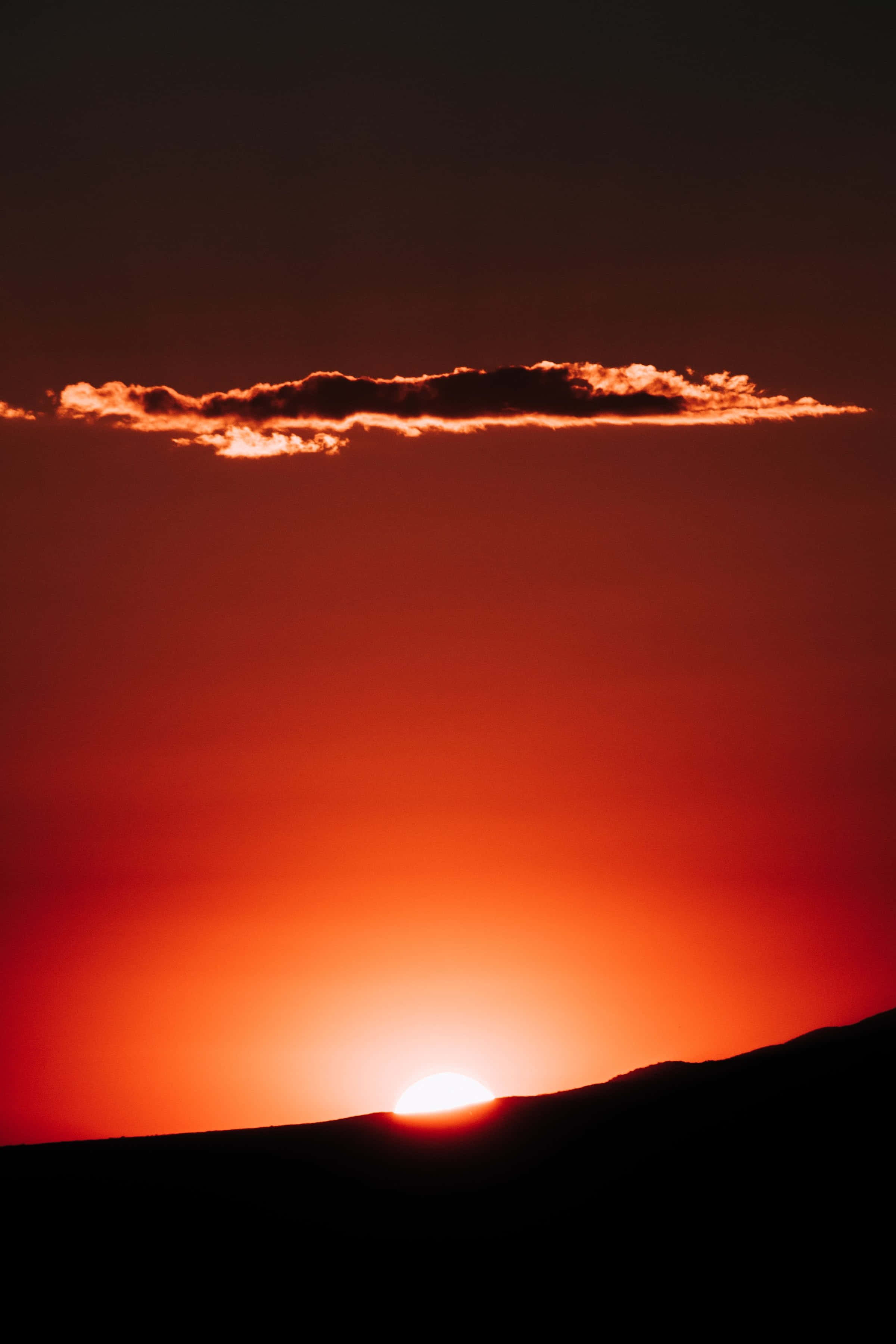 日落 桌布 壁紙 — 熾熱的夕陽將天空染成深橙色，太陽沉入黑暗的山脊之下，上方有一朵發光的雲。