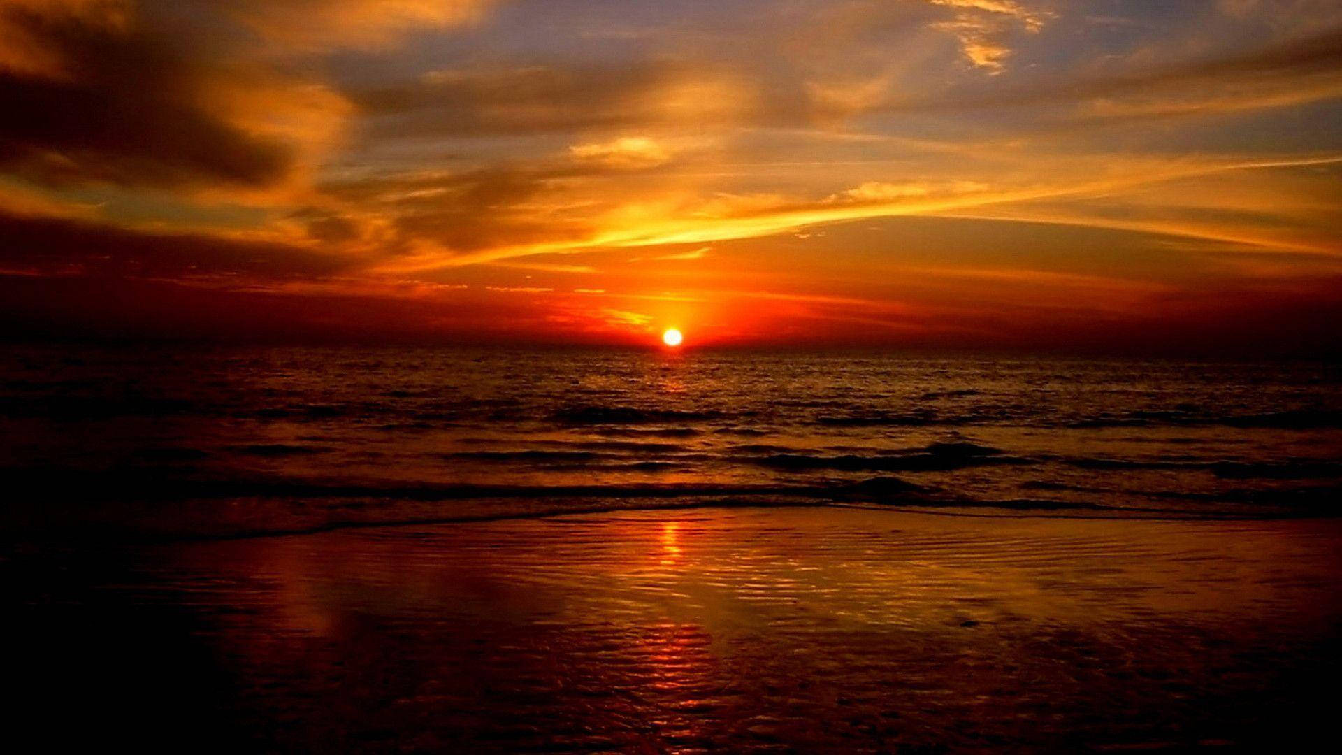 日落 桌布 壁紙 — 熾熱的夕陽將海洋和天空染成鮮豔的橙色、紅色和金色色調，海浪輕輕拍打著海岸。