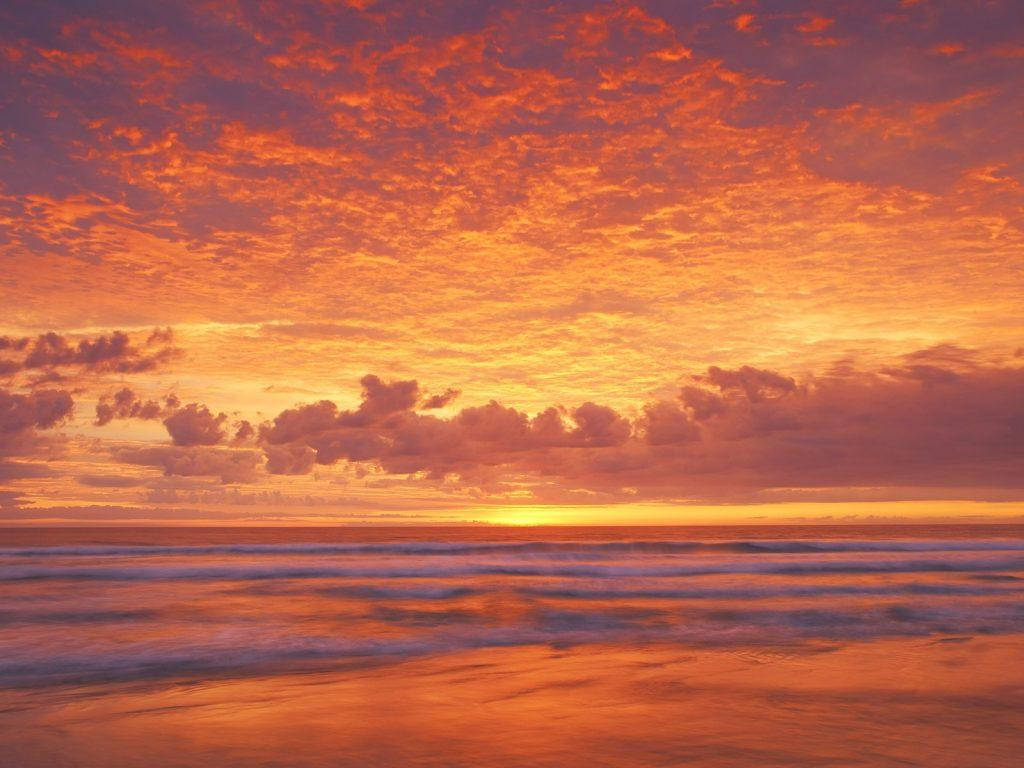 太陽 桌布 壁紙 — 熾熱的夕陽將海洋和天空染成鮮豔的橙色和紫色，海浪輕輕拍打著海岸。