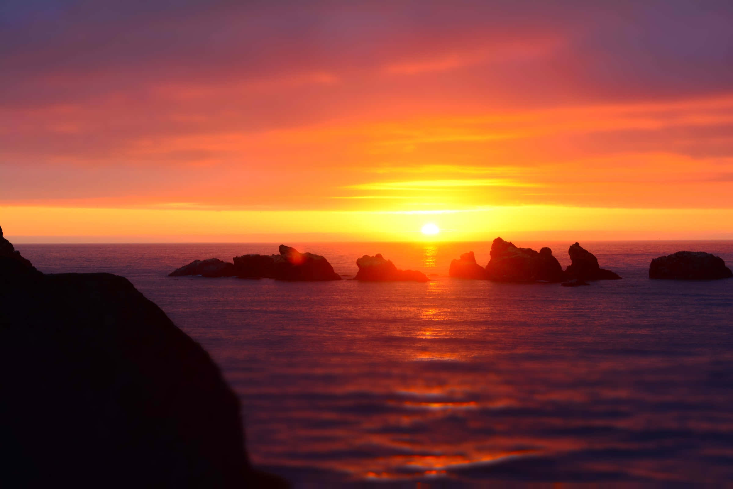 Sunset View 桌布 壁紙 — 熾熱的夕陽將海洋染成橙色和紫色，剪影般的岩石矗立在發光的地平線上。